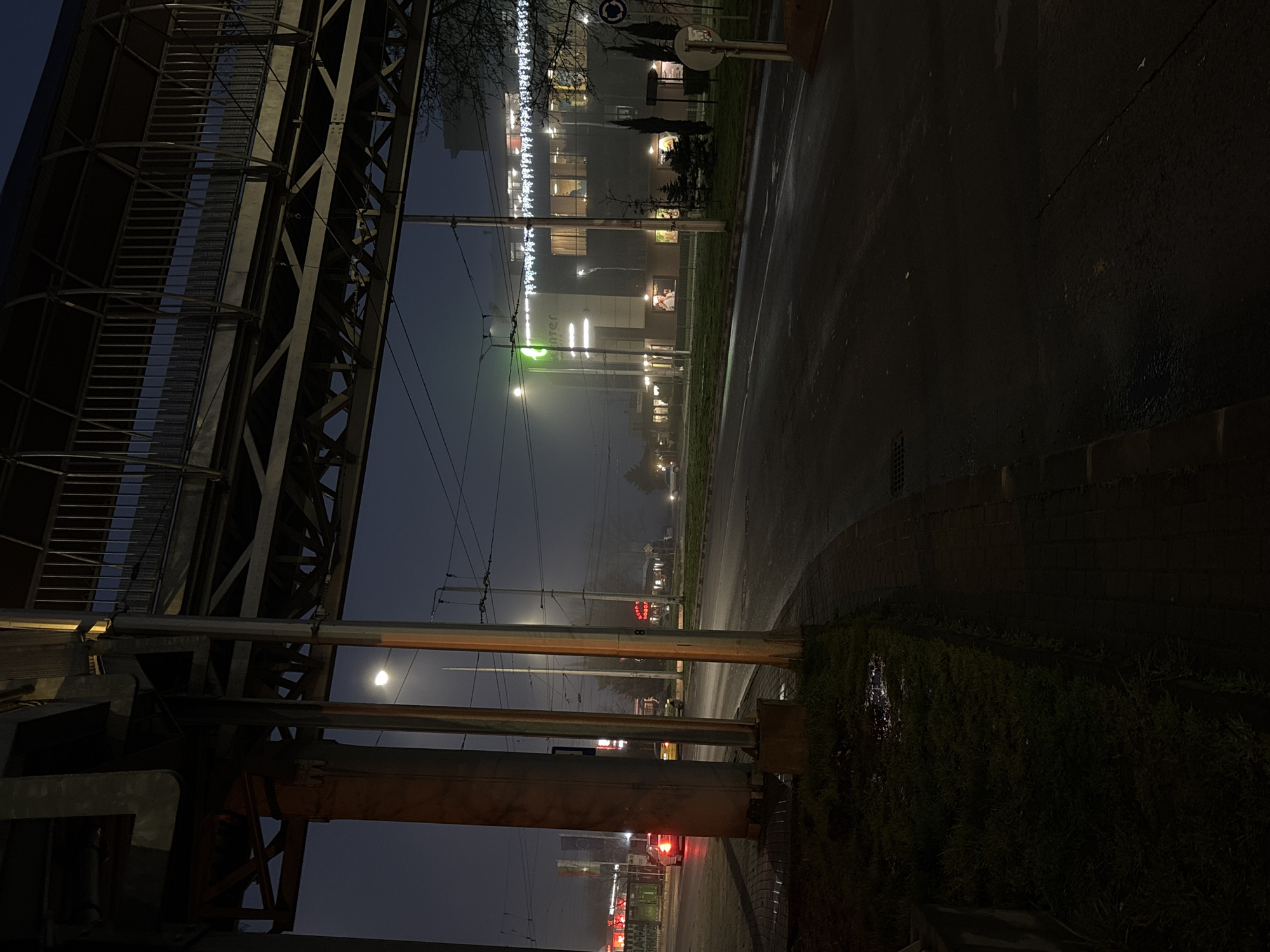 The image shows a night scene in Varna, Bulgaria. An elevated metal pedestrian bridge dominates the left side of the frame. Underneath the bridge, a traffic light is visible, indicating red. The view extends down a wet road, with illuminated buildings in the background. Street lights and overhead power lines are present. A low brick wall and vegetation run along the right edge of the frame, separating the road from a slightly higher area. The overall atmosphere is dark and likely rainy.