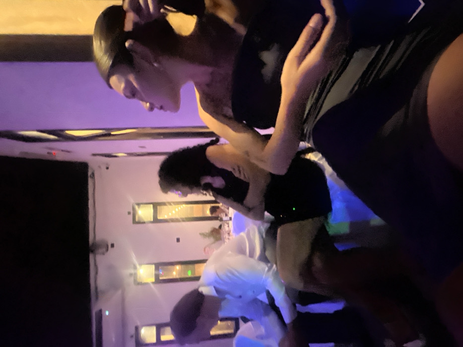 A low-angle shot shows multiple people gathered in what appears to be a banquet hall or party venue. The foreground features two women, one with slicked-back hair and a dark outfit, and another with curly hair, also in dark attire. Below them are other individuals, including one in a white shirt and light-colored trousers. Several rectangular windows with interior lighting punctuate the wall behind the group. The scene is bathed in a bluish-purple light, possibly from ambient lighting or spotlights, adding to the festive atmosphere.