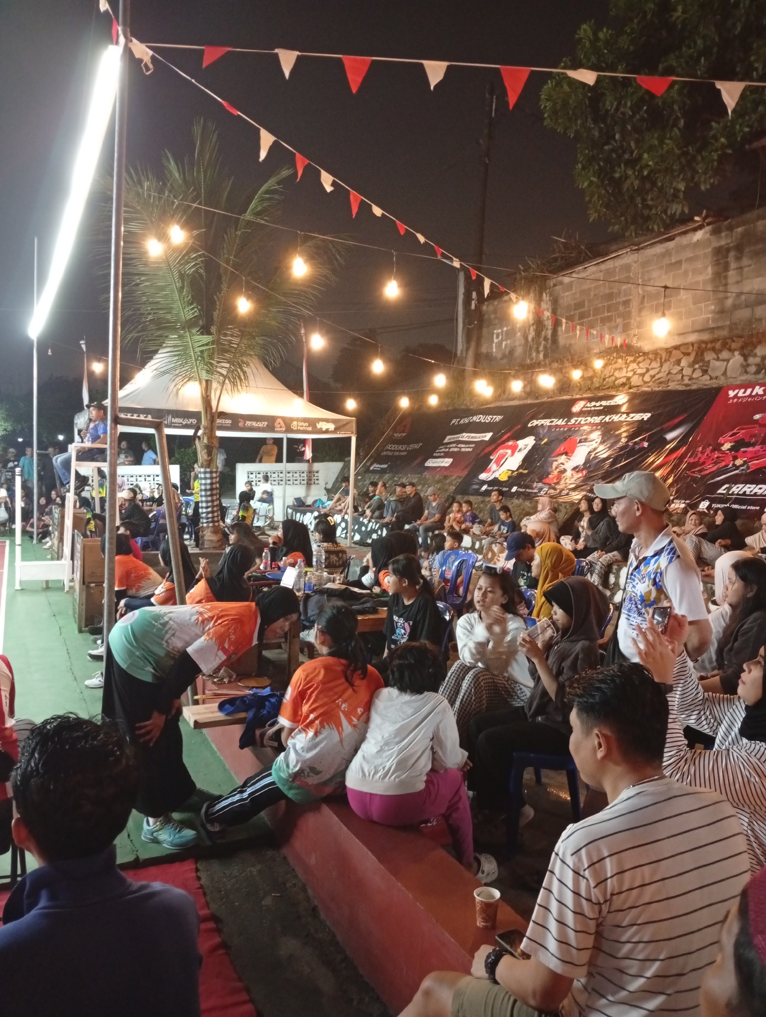 The image depicts an outdoor event at night, possibly a sporting event or a gathering, in Caringin, Indonesia. A crowd of people are seated on tiered benches, facing towards an unseen area. String lights and triangular red and white flags are strung overhead, creating a festive atmosphere. In the background, a large banner is displayed, featuring text in Indonesian and English, advertising "OFFICIAL STORE KHAZER" and "YUKIKU OFFICIAL STORE," suggesting a sponsorship or promotional event. A white tent is also visible, with a palm tree to its left. People of various ages are present, some dressed in casual clothing, while others wear colorful shirts. Some individuals are looking towards the direction of the event, while others are engaged in conversation or looking at their phones. The ground appears to be a court with green and red surfacing.