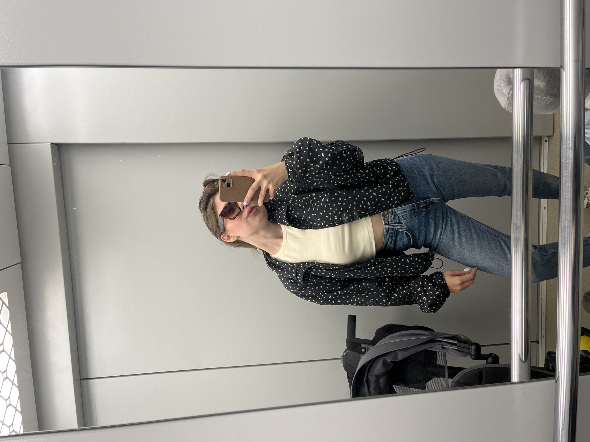 A young woman stands in an elevator, taking a selfie in the mirror. She is wearing a black patterned jacket over a cream-colored crop top and blue jeans. She has on large, square-framed sunglasses and her hair is pulled back in a bun. In the background, a stroller is visible, along with the plain gray walls of the elevator. There is no indication this scene is in Chaikovsky, Russia.