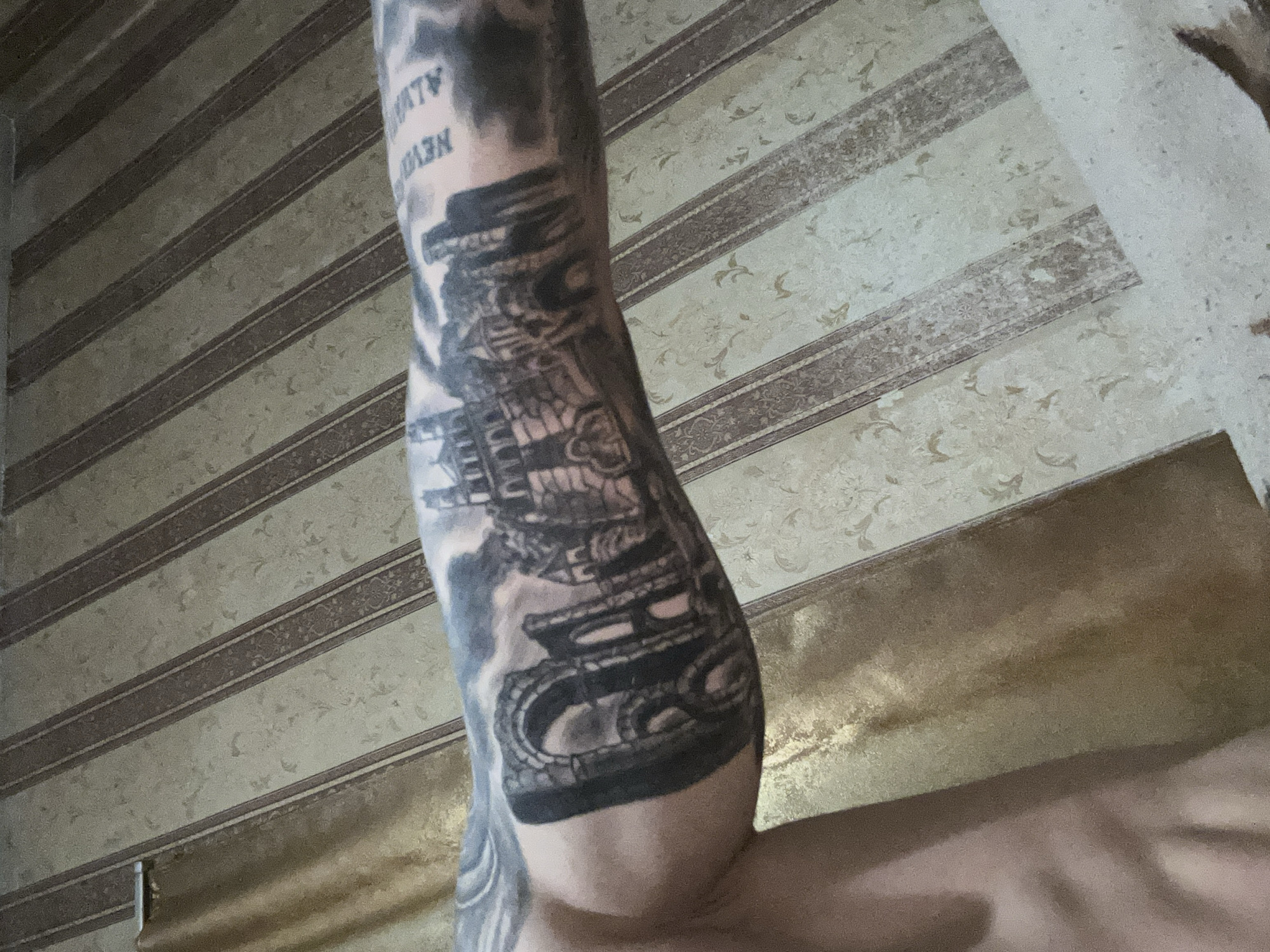 The image shows a person's arm with a black and grey tattoo on it, possibly a sleeve tattoo, featuring a complex design that includes architectural elements and perhaps text. The background is a patterned wallpaper. The location, Kalchevo, Bulgaria, is not ascertainable from the image.