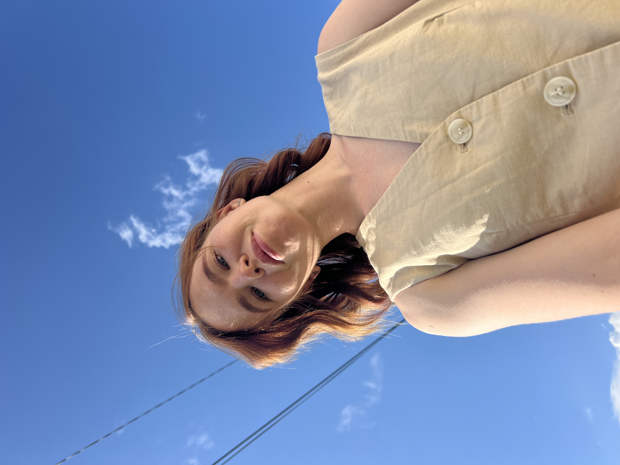 A young woman with red hair and freckles is pictured from a low angle, looking up at the camera with a slight smile. She is wearing a light-colored, sleeveless vest with two round buttons. The background is a clear blue sky with a few wispy white clouds and two parallel power lines running diagonally across the upper left quadrant of the frame. The lighting suggests it is daytime, possibly late afternoon due to the angle of the light casting shadows on her face. The image appears to have been taken outdoors, likely in a city setting given the presence of power lines. There is no identifiable information in the image to suggest a specific location in Chaikovsky, Russia.