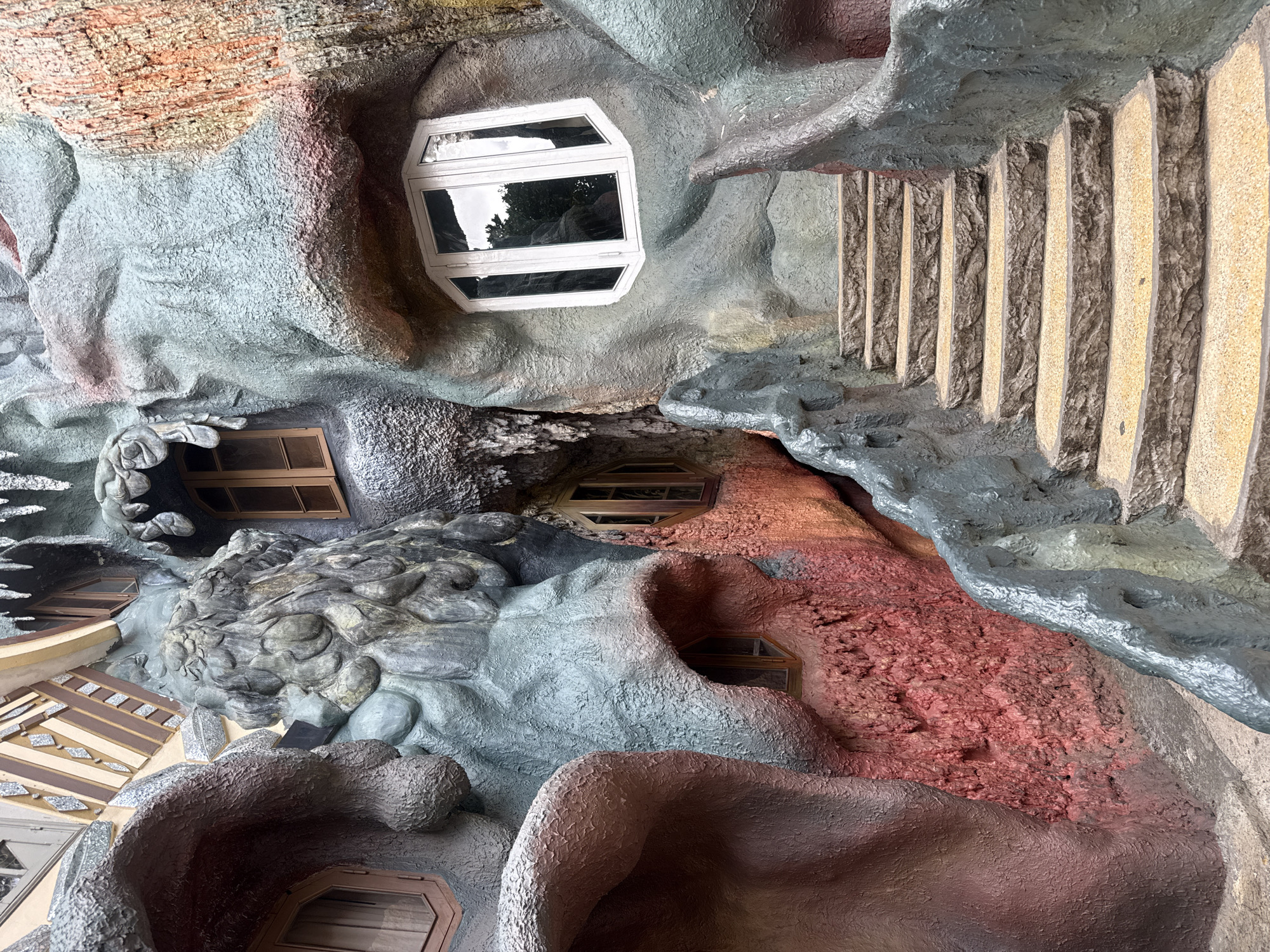 The image shows a section of the Crazy House in Da Lat, Vietnam. It features an outdoor staircase made of rough-hewn stone steps with yellow aggregate surfaces, flanked by organically shaped, textured walls. These walls are painted in mottled shades of blue-gray, red, and a sandy orange-brown, resembling natural rock formations or even monstrous organic structures. Several uniquely shaped windows are visible on the walls, some with wooden frames and others with white frames, reflecting the sky and foliage. The architecture is surreal and fantastical, creating a whimsical and slightly eerie atmosphere. The overall impression is of an otherworldly, sculpted environment.