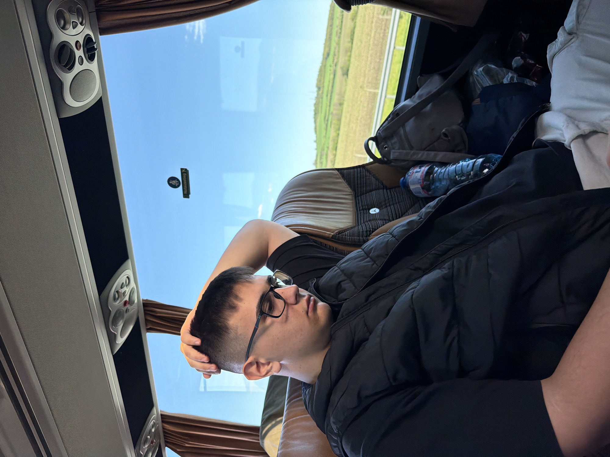A young man with short dark hair and glasses is reclining in a bus seat. He is wearing a black t-shirt and a black vest. His right hand rests on his head. A water bottle and a grey backpack are visible on the seat beside him. The bus window shows a view of a green field and a highway. Overhead compartments and curtains are visible. A sticker with a symbol and the word "Nooduitgang" (Emergency Exit) are also seen on the window. The exact location in Yazdach, Bulgaria is not discernable from the image.