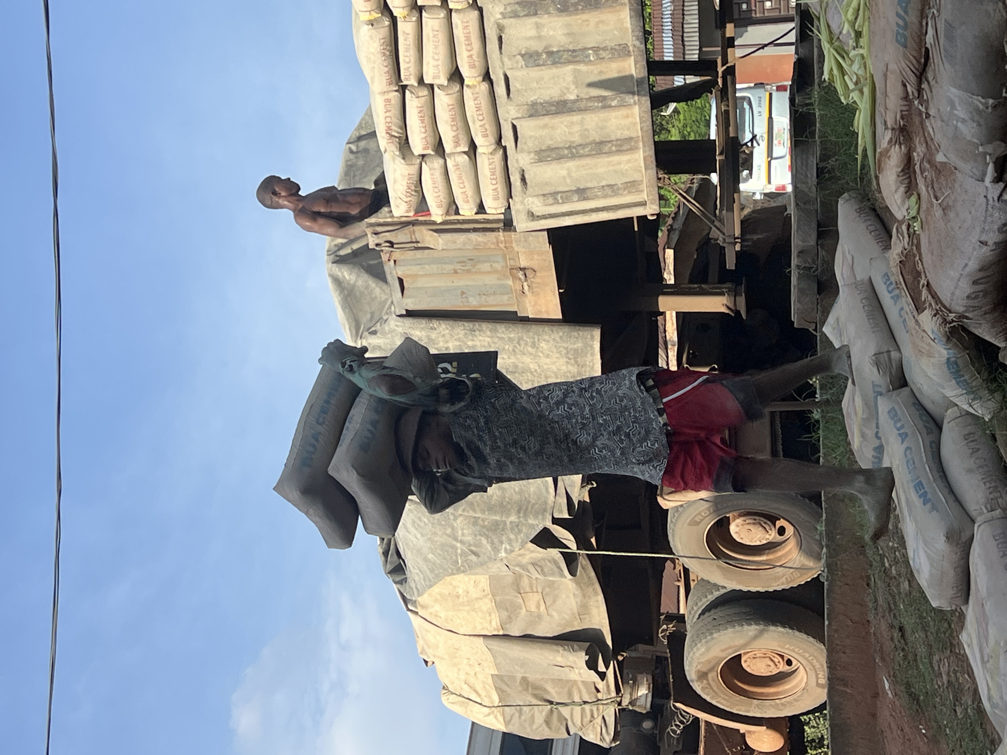 In Nkpor, Nigeria, a young boy carries a stack of cement bags on his head, his bare feet on other bags. He wears a patterned shirt and red shorts, with a tarp-covered truck loaded with more cement bags behind him. Another shirtless man stands on the truck bed. The scene is set against a blue sky with a power line overhead, and industrial elements like large truck tires are visible.