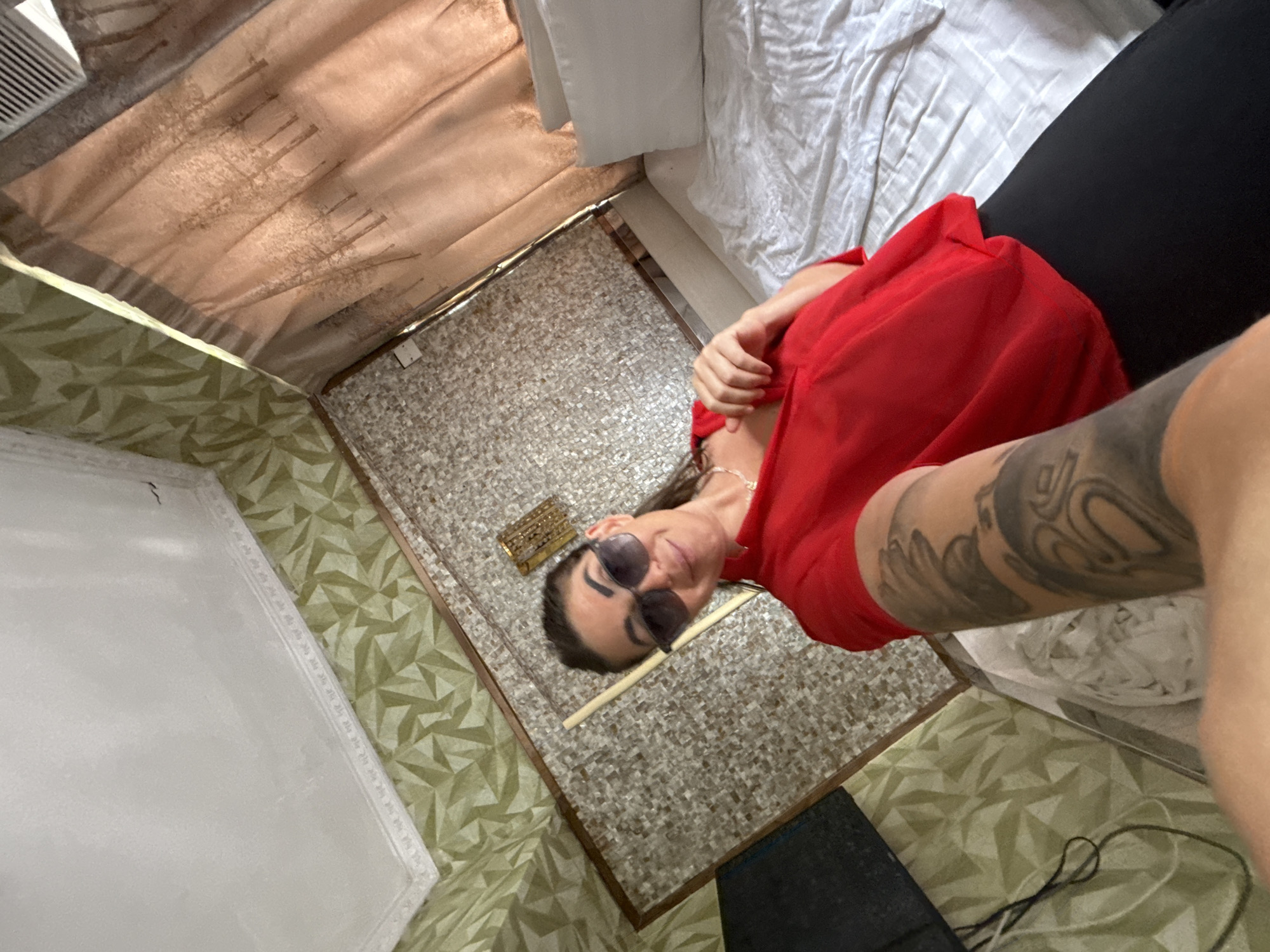 A woman with dark hair, wearing sunglasses, a red top, and black pants, is taking a selfie in a bedroom. She has tattoos on her left arm. The room has patterned wallpaper, a textured wall panel, a bed with white linens and pillows, and curtains with a tree motif. An air conditioning unit is visible in the upper right corner. The overall impression is of a casual indoor setting.