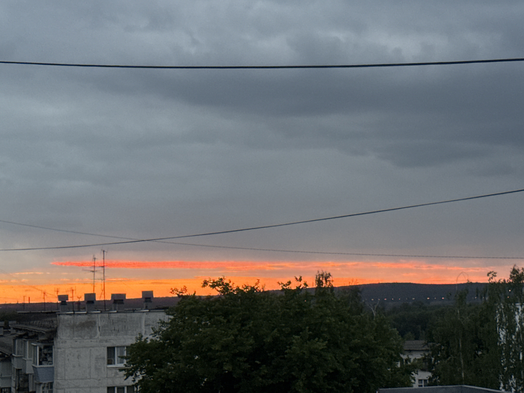 The image shows a dusk sky with a vibrant orange band stretching across the horizon, indicative of a sunset or sunrise. Above this colorful band, the sky is a muted gray with clouds. In the foreground, the rooftops of apartment buildings are visible, some with television antennas. A large, dark green tree partially obscures the view of the buildings and the horizon. Power lines are stretched across the upper portion of the image. The scene appears to be an urban environment, likely within the city of Chaikovsky, Russia, given the architectural style of the buildings. There are no people or animals visible in the frame, and no active events are taking place. The overall atmosphere is one of calm and the end of the day.