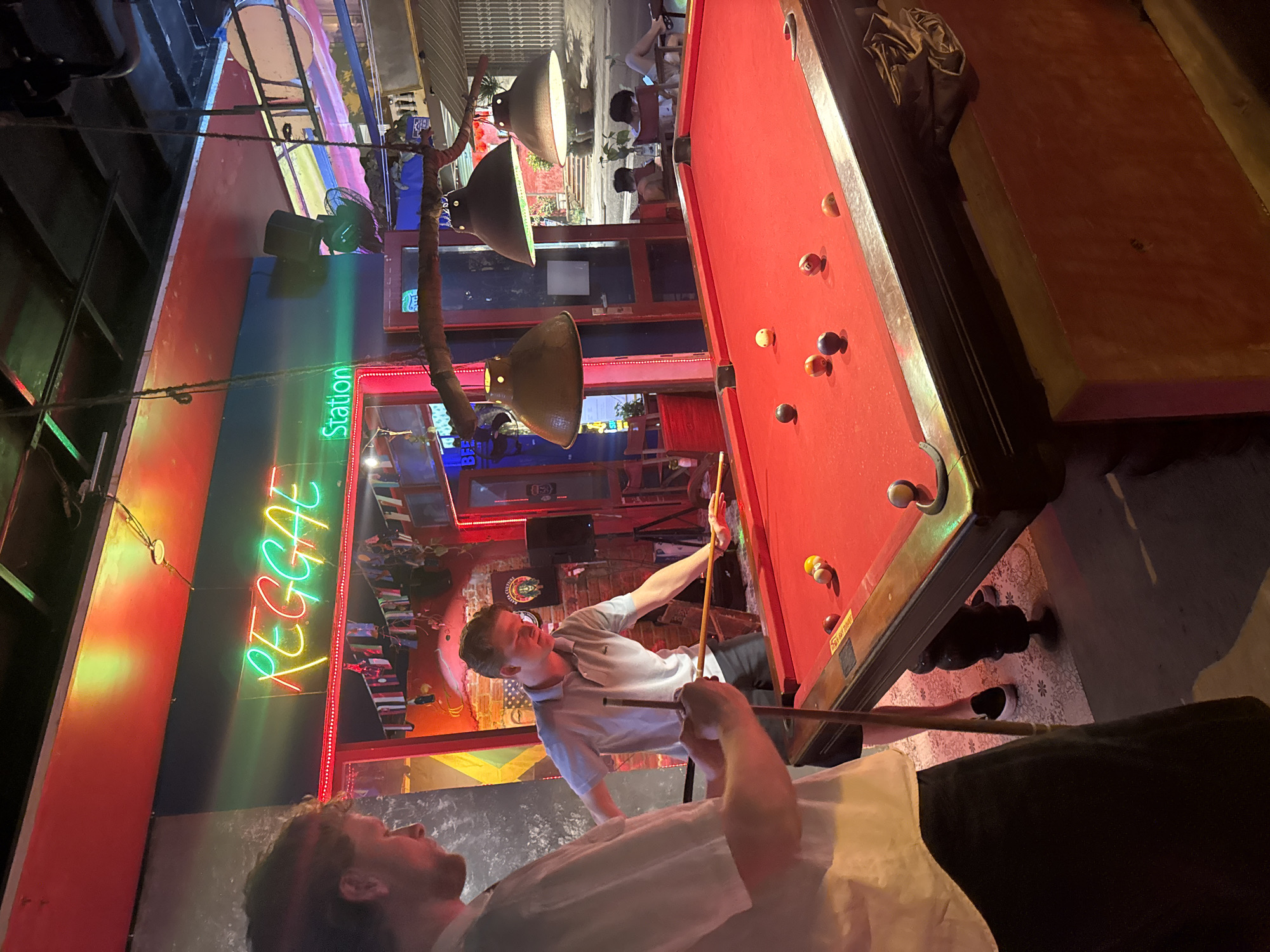 The image captures an indoor scene at what appears to be a bar or entertainment venue in Đà Nẵng, Vietnam, based on the "Reggae Station" neon sign visible on the left. Two men are in the foreground. One is standing and is in the process of lining up a shot on a pool table with red felt. The second man is positioned behind and to the left, facing towards the pool table. Patrons can be seen sitting at tables in the background, beyond the open front of the establishment. Lighting fixtures hang from the ceiling, illuminating the scene. The pool table is surrounded by a decorative tiled flooring and is located near the open front of the establishment, offering a view of the street beyond.