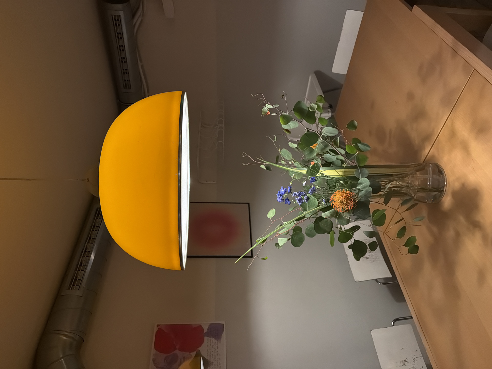 The image depicts an indoor scene within a modern establishment, identified as Kafe Zaokret at Nikole Spasića 3, Belgrade, Serbia. A large, rectangular light-brown wooden table occupies the central and lower portions of the frame. On the table, a clear glass vase holds a multi-component floral arrangement featuring broad green eucalyptus leaves, clusters of small blue flowers, slender green stalks, and a distinctive orange pincushion protea. Several white chairs with metal frames are positioned around the table, their backs partially visible and showing signs of wear.
Overhead, a prominent, inverted yellow dome-shaped pendant lamp is suspended, serving as the primary light source and casting a warm glow. Above the lamp, silver-colored exposed HVAC ductwork runs horizontally across the light-colored ceiling. The walls are painted a light gray hue. On the left wall, two framed artworks are visible: one with abstract red and yellow shapes and text, and another with a soft red-pink gradient circular form. In the upper right background, a white utility sink or basin is partially visible, and above it, a wall-mounted rack holds several white clothes hangers. The scene is static; no people are present, and no actions are depicted.