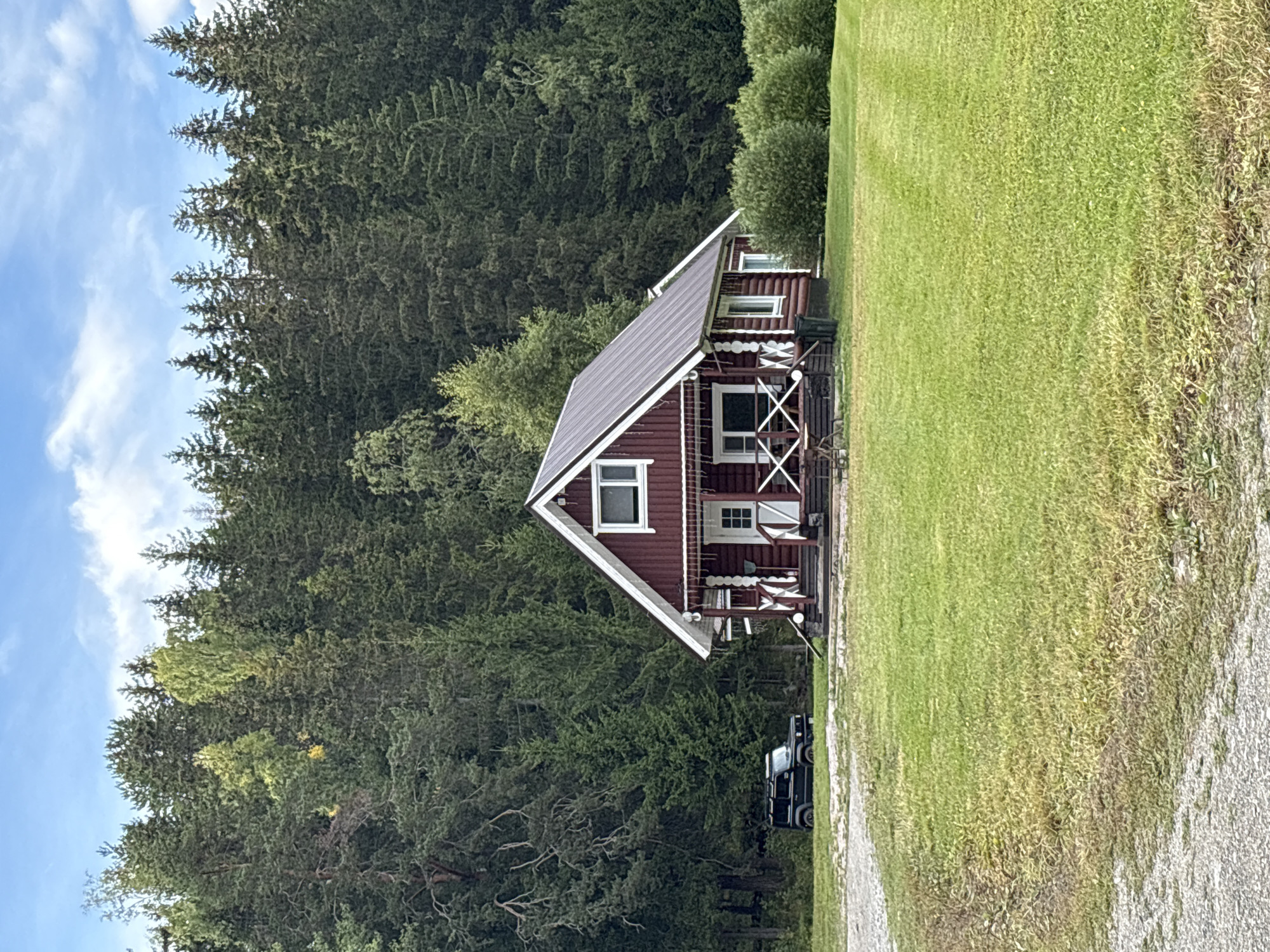 The image depicts a dark red log cabin with a metal roof, set in a grassy clearing. The cabin has white trim around the windows and doors, and a covered porch with white railings. A black SUV is parked to the left of the cabin. The background is dominated by a dense forest of pine trees. The sky is blue with scattered white clouds. The location is specified as Rautalakhti, Russia.