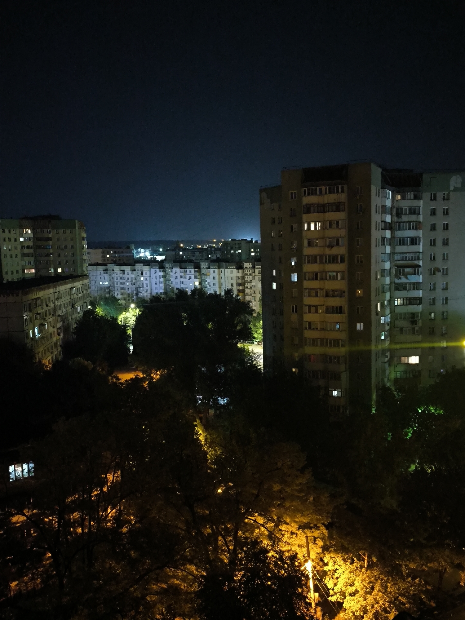 The image depicts a nighttime view of a dense urban residential area in Chișinău, Moldova. In the foreground, dark silhouettes of trees obscure much of the lower portion of the frame, their leaves illuminated by the warm glow of streetlights and artificial lighting from buildings.
Dominating the mid-ground and background are several multi-story apartment buildings, characteristic of Soviet-era architecture, with numerous windows glowing with interior lights, suggesting occupancy. The buildings vary in height and facade, some appearing older and weathered with exposed concrete, while others show signs of renovation. A distinct, tall, and more modern-looking apartment building stands prominently on the right side of the frame, its facade detailed with many windows and balconies.
Further in the distance, more apartment blocks are visible, their lights forming a diffused urban glow against the dark night sky. The overall impression is that of a typical residential neighborhood in a city at night, with the trees in the foreground adding a natural element to the man-made environment. There are no visible people or specific activities depicted. The scene is calm and static, conveying the quiet of a city after dark.