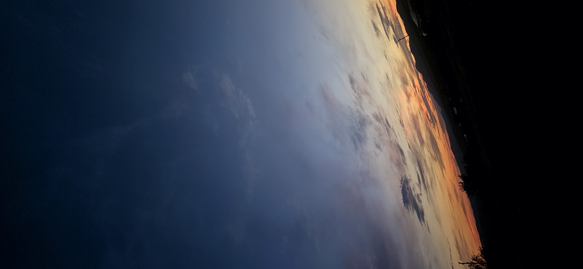The image is a vertical, wide-angle shot of a sunset over Momin Prohod, Bulgaria. The sky transitions from a deep blue at the top to lighter shades of blue, pink, and orange near the horizon. Wispy clouds are scattered across the sky, catching the sunset colors. In the foreground, silhouetted trees and hills are visible against the vibrant sky. The landscape appears rural, with distant mountains forming the horizon line. There are no people or animals visible in the image. The overall mood is tranquil and serene.