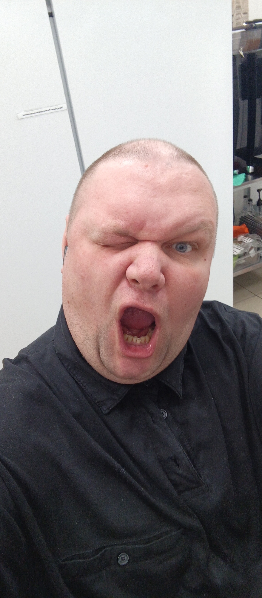 A medium shot, eye-level, selfie-style photograph captures a bald, fair-skinned man with a large head and light blue eyes, winking with his left eye and his mouth wide open in a yell or scream. He is wearing a black collared shirt. A thin, grey vertical bar runs along the left side of the frame, partially obscuring the white wall behind him. A small, white rectangular sticker with black text is affixed to the wall, visible to the left of the man's head. The background is predominantly a plain white wall, with a blurred section of shelves and items to the right, suggesting an indoor environment. The man's face takes up most of the frame, with the focus on his expressive facial features and open mouth. There is no indication of the city of Bugry, Russia, within the image itself.