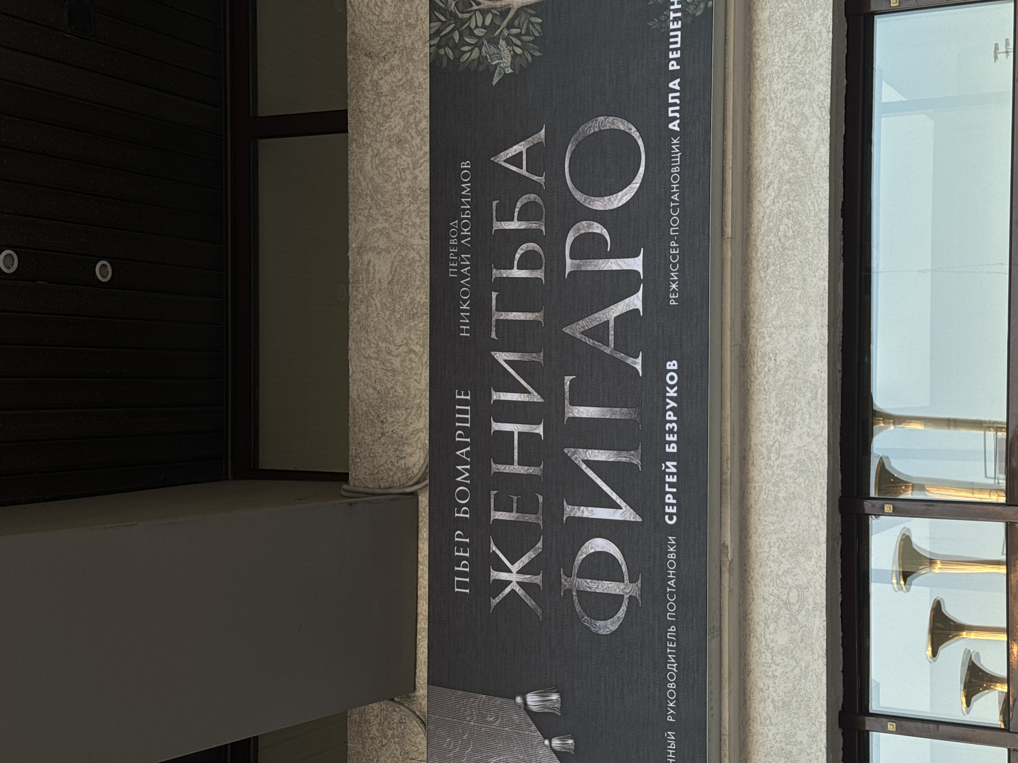 The image shows an outdoor sign in Moscow, Russia advertising a production of "The Marriage of Figaro." The sign lists the playwright as Pierre Beaumarchais, the translator as Nikolai Lyubimov, and the director as Sergei Bezrukov. The director-producer is named as Alla Reshetn. A set of golden-colored horns are visible behind the sign in a window.