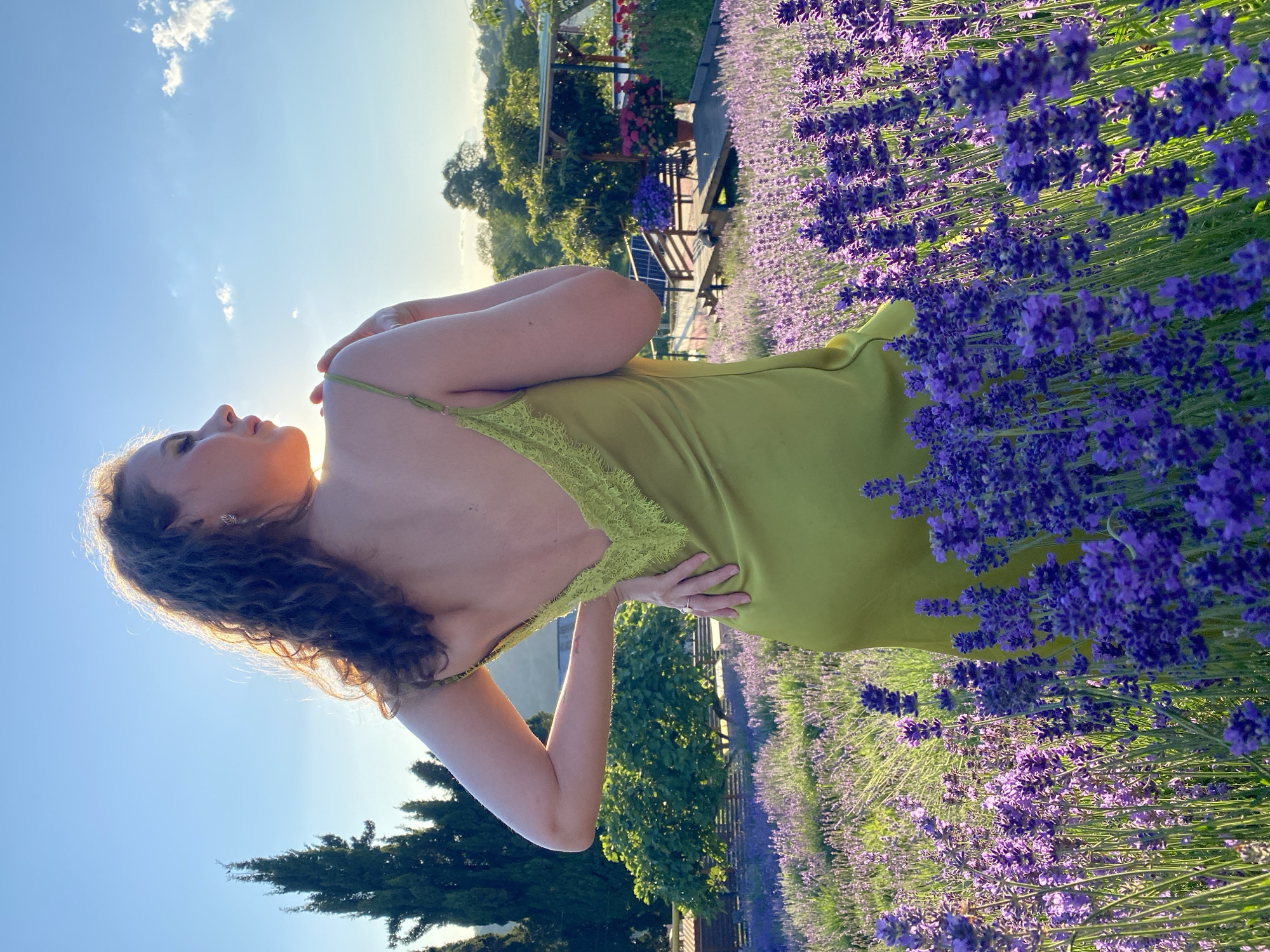 A woman with curly brown hair, wearing a lime green slip dress, stands facing away from the camera in a field of lavender. She has one hand on her hip and the other touching her shoulder. The sun is setting behind her, casting a warm glow. In the background, there are trees, a wooden structure, and a clear sky with scattered clouds. The location is Verkhneye Uchdere, Russia.