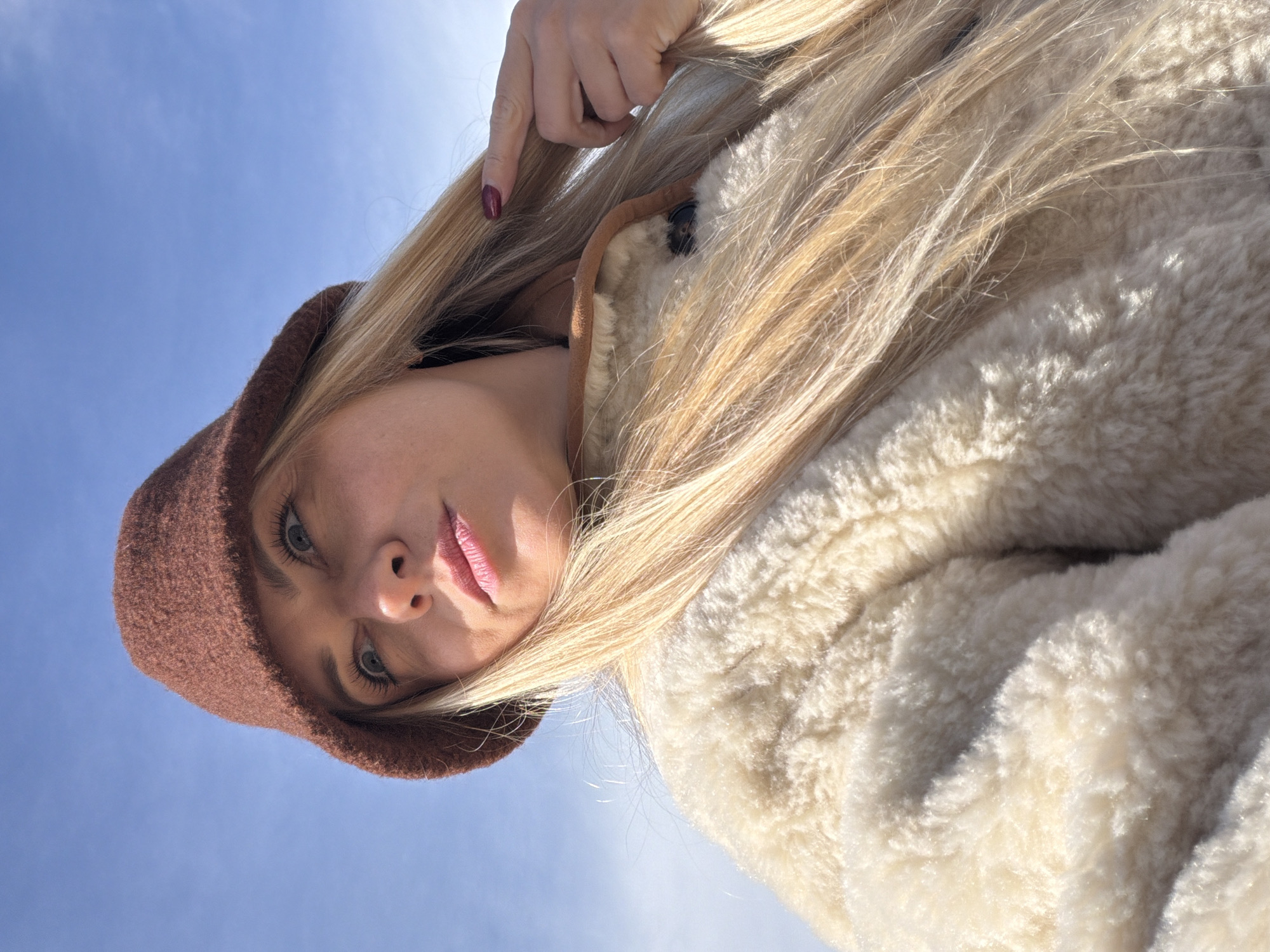 A woman with blonde hair and blue eyes is seen from a low angle perspective, looking down at the camera. She is wearing a brown knit hat and a cream-colored fur coat. Her right hand is visible near her hair, with a red-polished fingernail showing. The background is a light blue sky. The location is not discernible as Votkinsk, Russia, from the image.