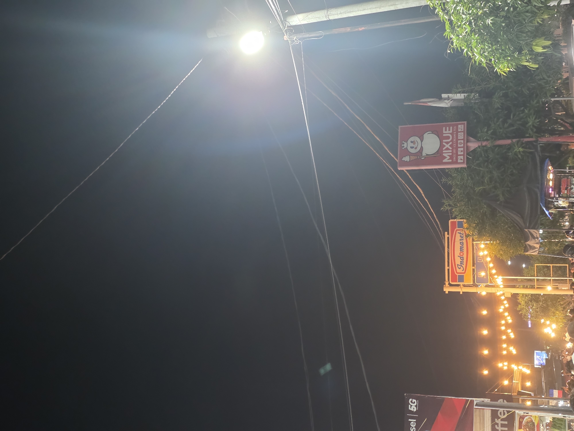 This is a night scene in the city of Bireuen, Indonesia. A street light is prominently visible on the right side of the image, casting a bright glow. Several power lines crisscross the dark sky. In the background, there are signs for businesses, including "Indomaret" and "Mixue". String lights illuminate a pathway or market area. There are also trees and the tops of various structures, suggesting an urban or semi-urban environment. In the lower part of the image, a crowd of people is gathered, indicating a public space, possibly a market or event. A faint contrail from an aircraft is visible in the upper portion of the sky.