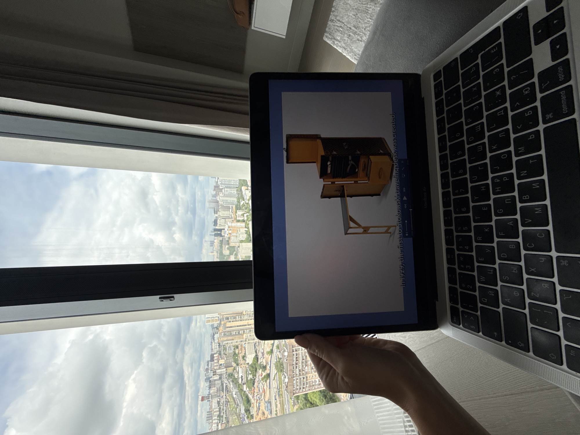 The image captures a person's left hand holding a laptop in front of a large window, offering a panoramic view of a city. The laptop is identified as a "MacBook Air" and features a keyboard with both Latin and Cyrillic characters.
The screen of the laptop displays what appears to be a video or presentation. A prominent image on the screen shows a vintage-style, open wooden trunk or "traveling desk." Below this image, text is visible that reads, "In 1929, the first to Stokowski traveling desk was created." Video playback controls, including play/pause buttons and a timeline indicating a duration (e.g., 0:01 / 7:30), are also present at the bottom of the screen.
Through the window, a vast urban landscape stretches under a cloudy sky. Numerous high-rise buildings, roads, and some construction sites are visible, characteristic of a modern city. The weather is overcast but bright, suggesting daytime. The setting is indoors, likely a high-floor apartment or office, given the extensive city view. The presence of the Cyrillic keyboard and the urban landscape are consistent with the provided location context of Khimki, Russia. The scene depicts a casual moment of media consumption on a laptop with a scenic city backdrop.