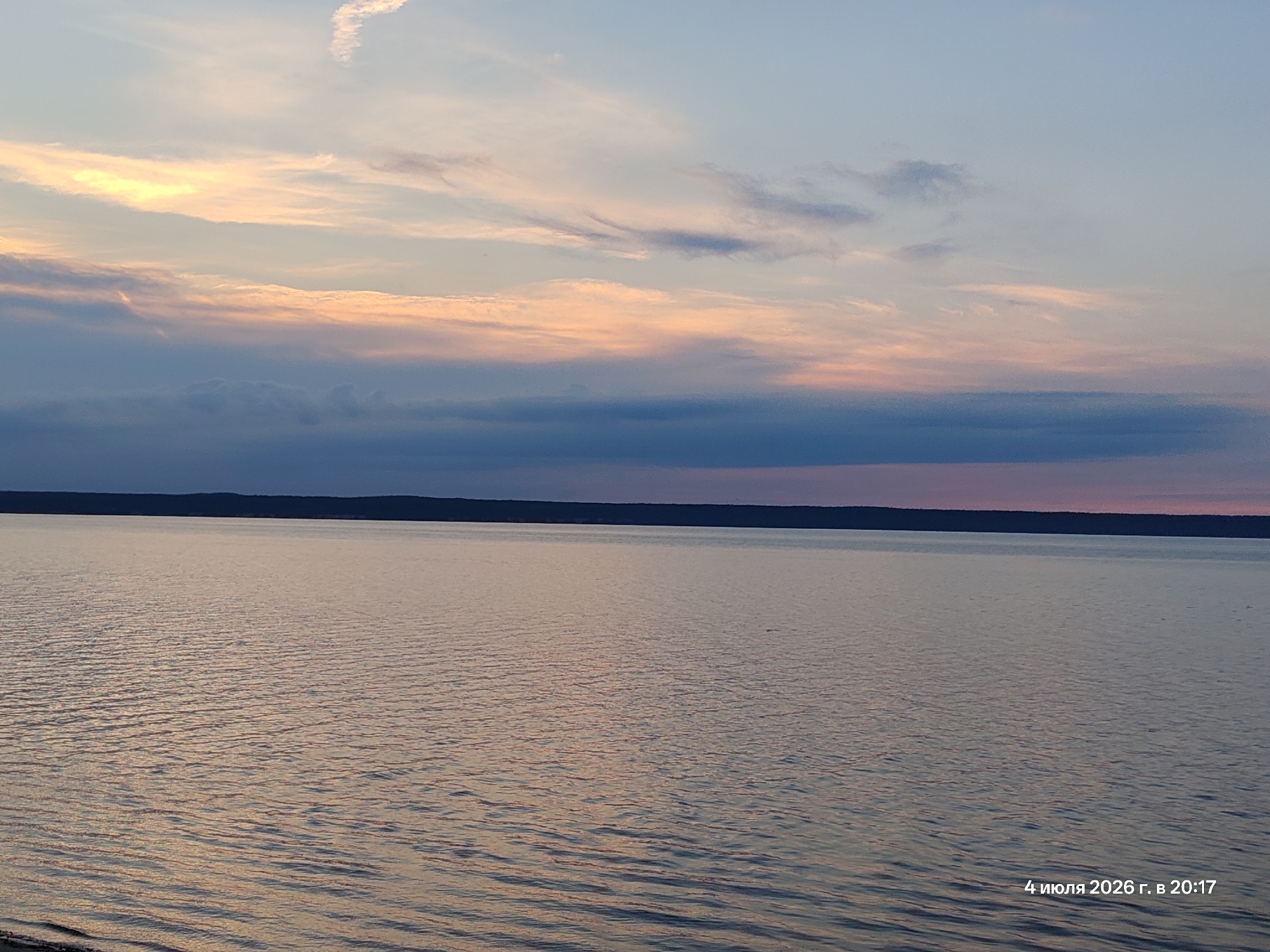 A wide, horizontal, eye-level shot captures a serene sunset over a large body of water. The sky is a canvas of soft pastel colors, with streaks of yellow, pink, and blue clouds stretching across the horizon. A dark, silhouetted landmass is visible in the distance, forming a straight line across the image. The water's surface is calm, with gentle ripples reflecting the colors of the sky. The overall mood is peaceful and tranquil. The date "4 июля 2026 г. в 20:17" is visible in the bottom right corner, indicating the time and date the photograph was taken.