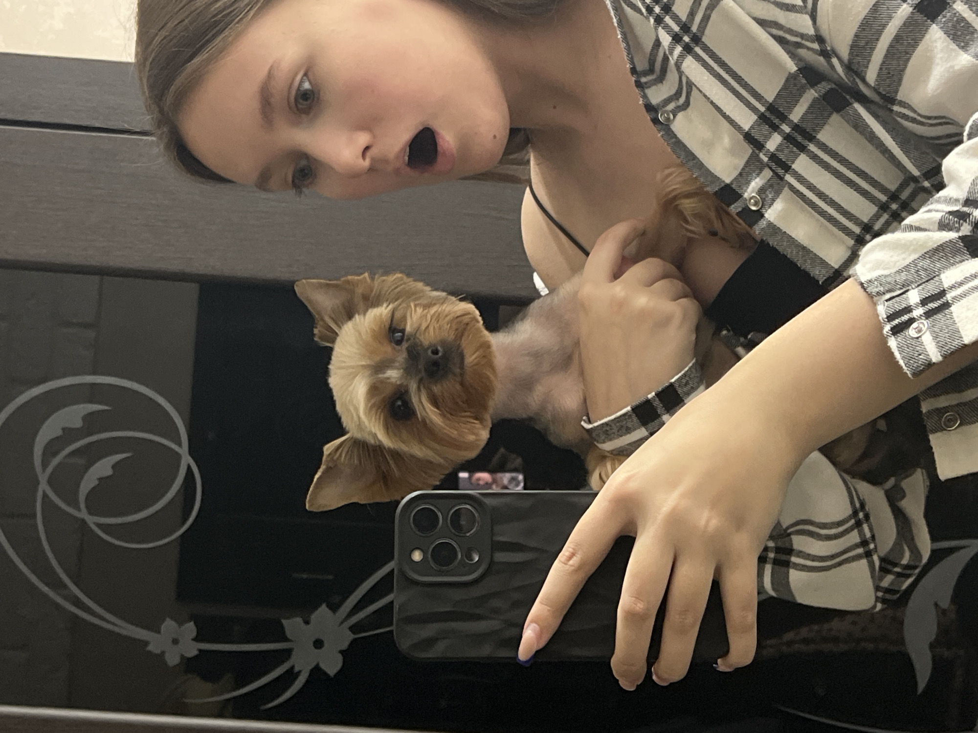 An angled, indoor photograph captures a young woman and a small dog reflected in a dark, glossy surface, likely a mirror or polished table, in Chaikovsky, Russia. The image is taken from above, looking down at the reflection.
The central figures are a young woman and a small, light brown dog, possibly a Yorkshire Terrier. The woman, with light brown hair and fair skin, is seen from the chest up, wearing a black and white plaid button-up shirt over a black top. Her mouth is open in a wide, surprised, or playful expression, and her eyes are wide, looking towards the viewer. She holds the dog close with her left hand, which is mostly out of frame. Her right hand, with a visible blue painted fingernail, holds a black smartphone with a textured case and three prominent camera lenses, indicating she is taking a mirror selfie or photographing the reflection.
The small dog is positioned above the smartphone, its face looking directly at the camera with an attentive expression. Its fur is well-groomed, and its ears are perked.
The reflective surface itself is dark and shiny, featuring white decorative etchings of swirling circles and floral patterns on the left side, visible beneath the woman's head and the dog. The background behind the reflection is indistinct and dark, suggesting interior elements like furniture or a wall. The overall mood of the image is playful and candid, capturing a moment between a pet owner and her dog.