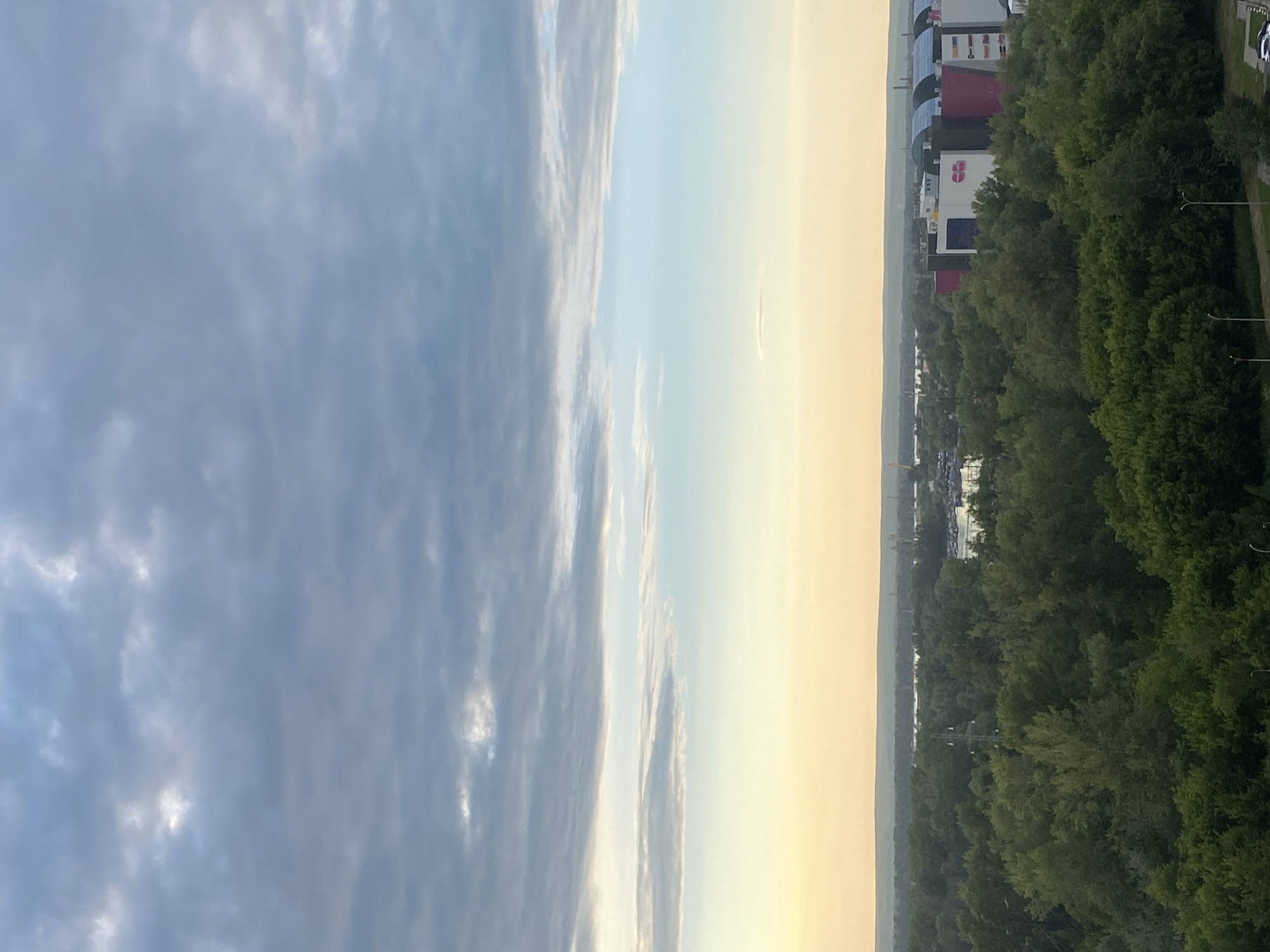 This is an aerial, high-angle, medium shot of a city skyline with a body of water in the background, likely during the golden hour due to the warm, soft lighting. The sky is filled with a mix of grey and white clouds, with a gradient of pale yellow and blue towards the horizon. A dense, green forest occupies the middle ground, obscuring much of the ground-level activity. In the upper right quadrant of the image, a cluster of modern buildings with a colorful, retail facade is visible, suggesting a commercial or shopping district. The buildings are distinct with curved, colorful roofs and large windows. A hint of a river or large lake runs horizontally across the landscape behind the forest. In the far distance, industrial elements such as cranes and smokestacks can be seen, indicating industrial activity. There are no people or specific actions captured in the frame, rendering the scene a static, environmental portrait of the city. The location is the city of Novokuybyshevsk, Russia.
