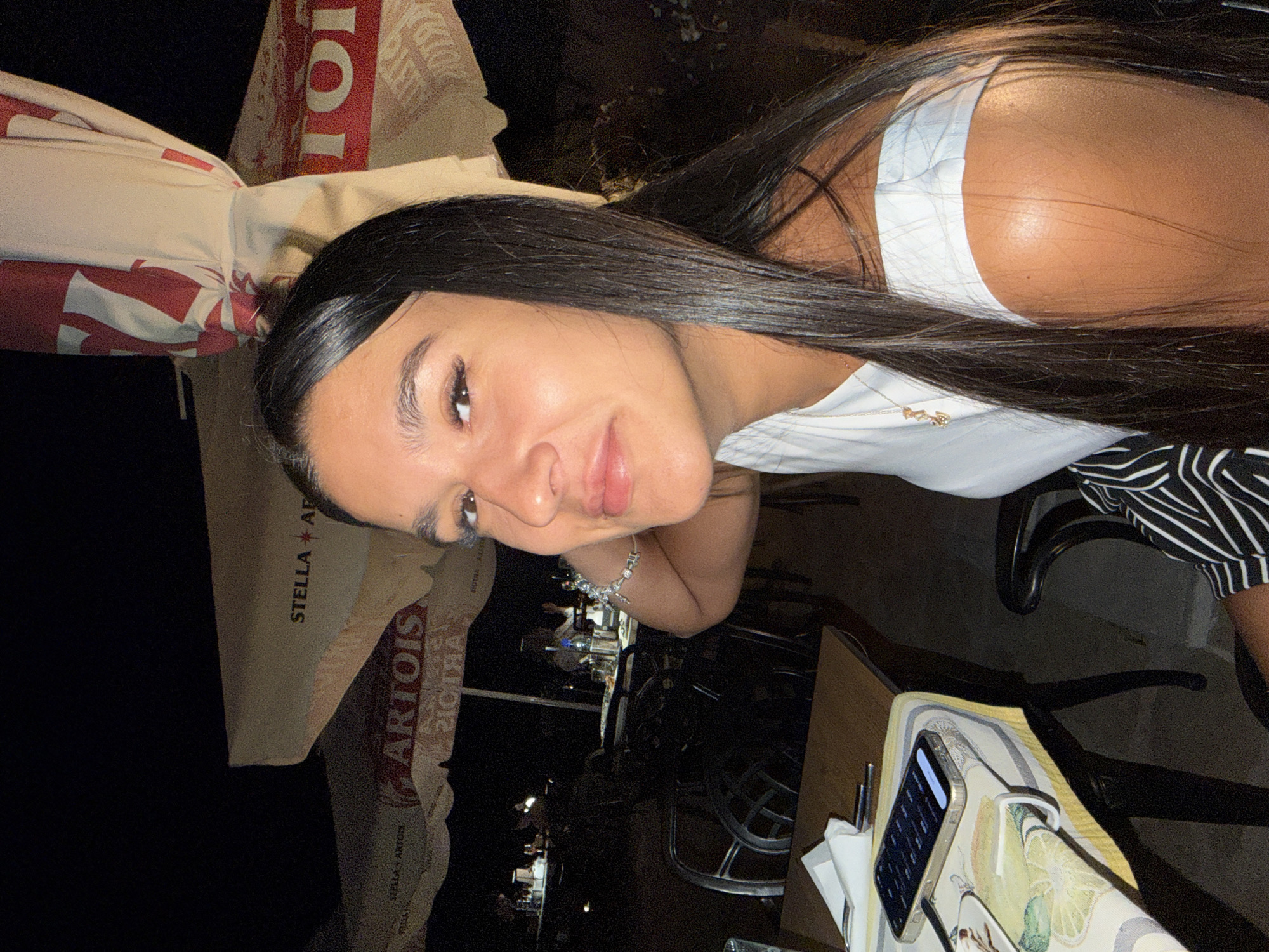 A young woman with long, dark hair is seated at a table outdoors at night. She is wearing a white off-the-shoulder top and a striped skirt. A silver bracelet adorns her wrist, and a delicate necklace hangs around her neck. In front of her, on the table, rests a smartphone. The background reveals umbrellas with logos for "STELLA ARTOIS," suggesting a bar or restaurant setting. The overall ambiance is casual and relaxed, likely in a European establishment. The scene is set in the historic town of Arbanassi, Bulgaria.