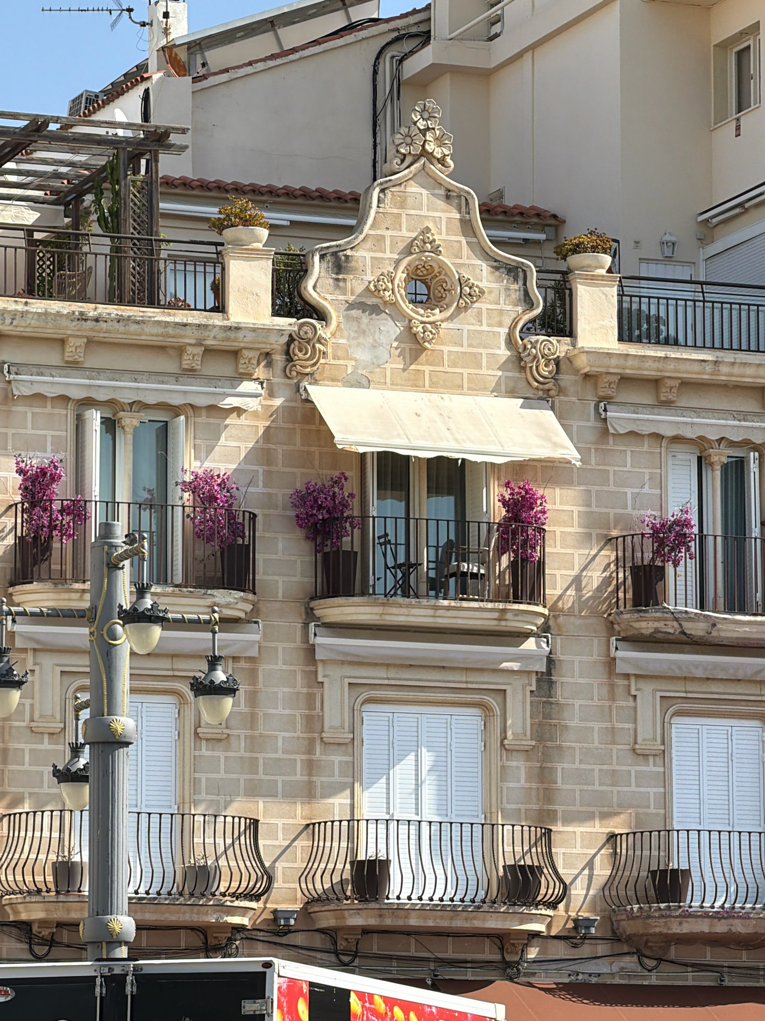 The image depicts a sun-drenched facade of a building in Sitges, Spain, characterized by its warm, sandy-colored brickwork and ornate architectural details. The building features multiple balconies with black wrought-iron railings, adorned with vibrant pink flowering plants in dark pots. Above the balconies, white awnings provide shade for windows. The upper levels of the building reveal more balconies and rooftop elements, including pergolas and potted plants. A prominent decorative pediment sits above the central upper-level window, featuring intricate stone carvings. A lamppost with multiple light fixtures stands in the foreground, adding to the street-level ambiance. The scene suggests a tranquil urban environment, likely a residential or commercial area.