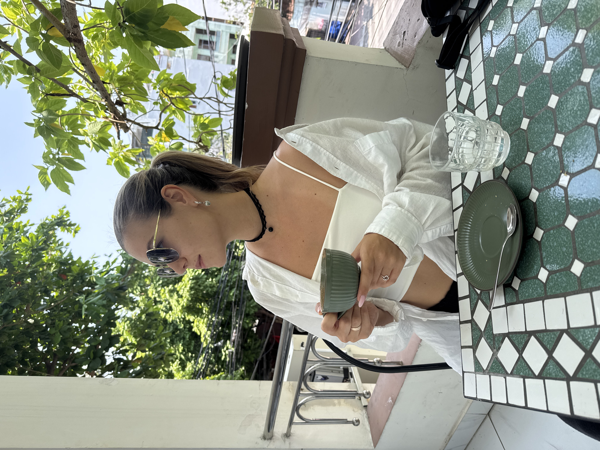 A woman is sitting at a table outdoors, likely on a balcony or patio, with lush green trees visible in the background. She wears sunglasses, a black choker necklace, a white top, and a light white button-up shirt. She's holding a small, green bowl in both hands. On the table is a patterned green and white mosaic tile design. Also visible are a clear glass, a green saucer with a spoon, and a black object partially visible at the top of the table. The location appears to be a balcony with lush green vegetation visible in the background in an unknown city in an unknown state.