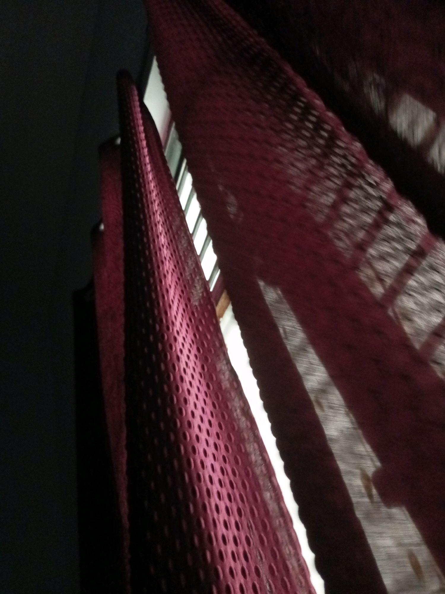 The image displays a close-up, low-angle view of red, perforated curtains indoors. The curtains are partially drawn, revealing vertical blinds behind them that are illuminated by an unseen light source, possibly sunlight, creating distinct diagonal streaks of light and shadow across the fabric. The texture of the curtains is a prominent feature, with a repeating pattern of circular perforations. The lighting creates a sense of depth and highlights the translucent nature of the material. The background is dark, with a portion of what appears to be a window frame visible to the right. The overall impression is one of a domestic interior setting, possibly a room in Mathura, India, where the interplay of light and shadow on the textiles is the primary focus. There are no people or overt actions depicted.