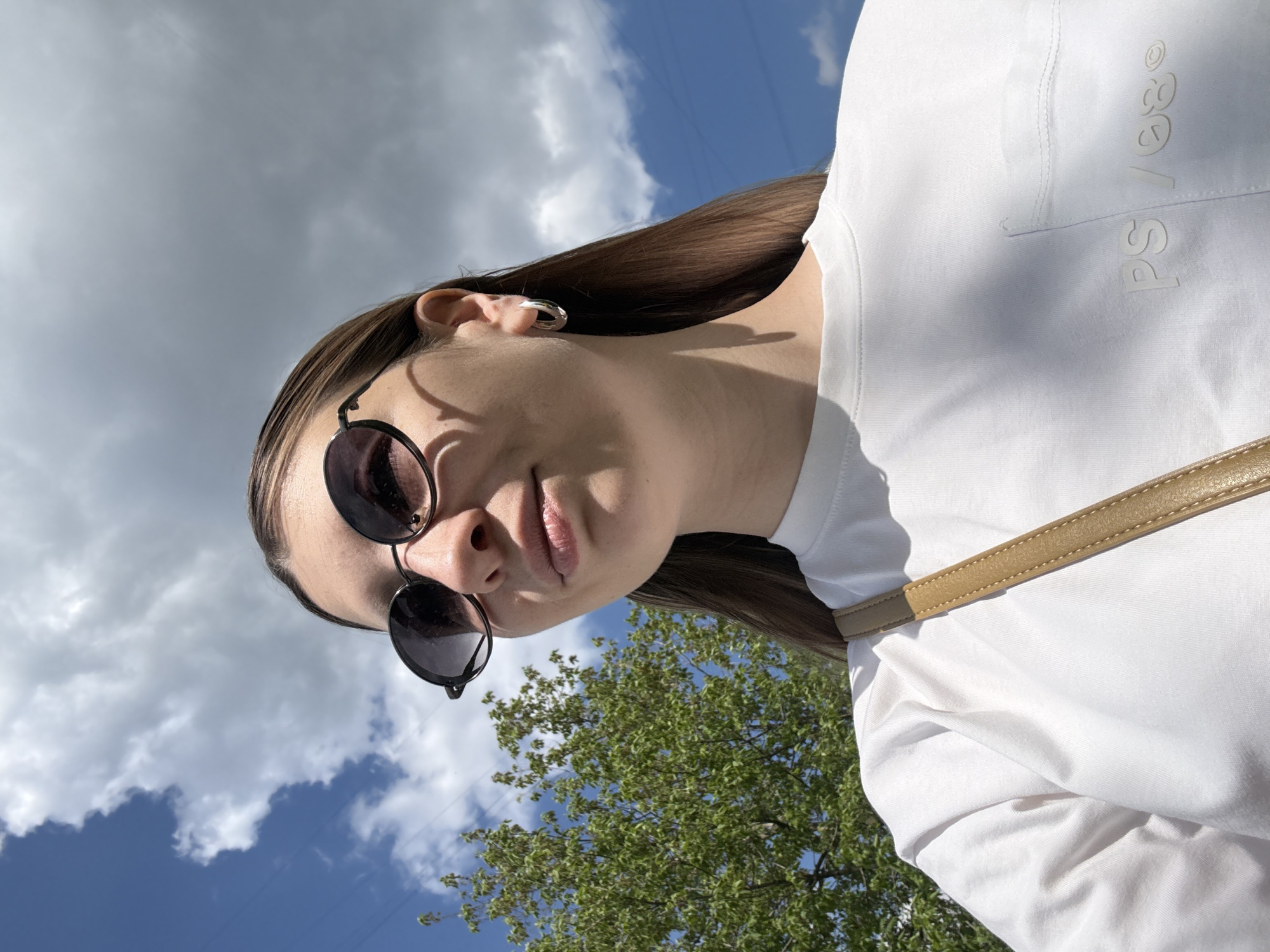 The image is a low-angle selfie of a woman wearing sunglasses and a white long-sleeved shirt. The background includes a blue sky with white clouds and a green tree. The text "PS/ses" is visible on the upper right portion of the shirt. A tan-colored strap, possibly of a bag, is also visible across the right side of the shirt. There is no indication of this photo's location being in Moscow, Russia.