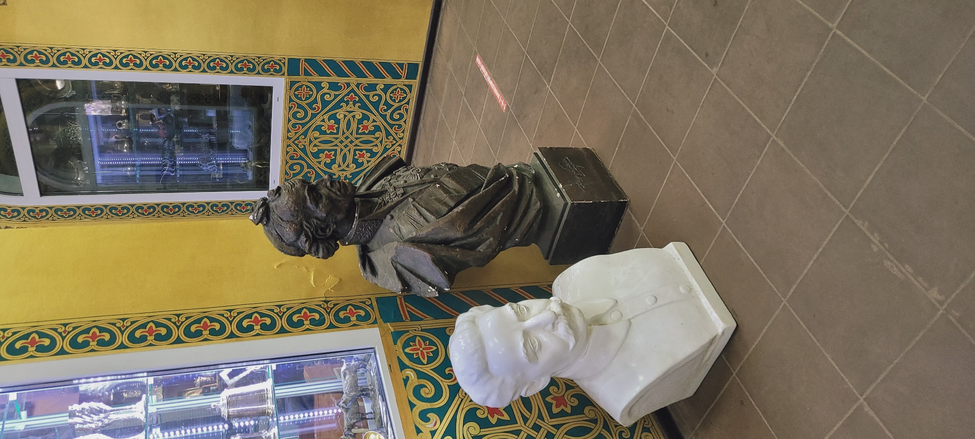The image captures an indoor scene with two busts prominently displayed against a backdrop of decorative elements and display cases. One bust, a bronze figure dressed in elaborate attire, stands on a dark-colored plinth. Next to it, a white bust depicting a man in a suit complements the display.
The background consists of a yellow wall adorned with an ornate, patterned frieze. Two illuminated display cases are visible, containing various objects. The floor is tiled in a dark, neutral tone. A red stripe marker is visible on the floor. The specific location within the city of Unknown, Unknown, cannot be identified from the image provided.