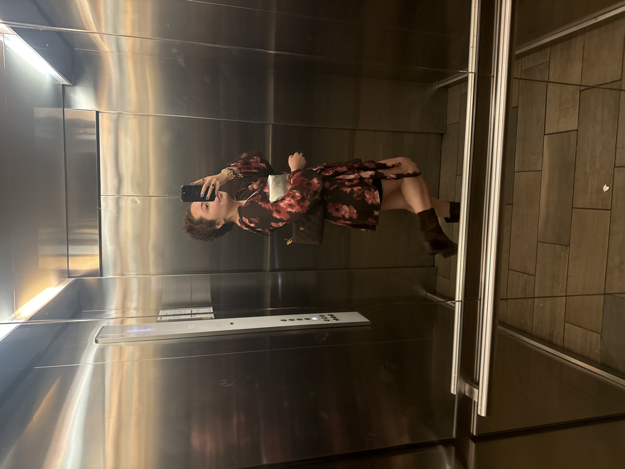 A woman with curly hair stands in an elevator, holding a black smartphone up in her left hand and taking a selfie. She's wearing a dark, floral-patterned dress and brown heeled boots, and she carries a brown purse on her left shoulder. The elevator has mirrored walls. Outside the elevator doors, the walls are covered in brown rectangular tiles. The location is possibly in Mexico City, Mexico.