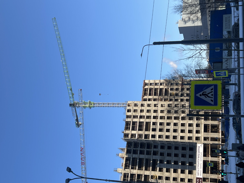 This image depicts a construction site on a clear, sunny winter day in Saint Petersburg, Russia, specifically identified as the vicinity of the SAAN PARK residential complex, likely near Bogatyryorsky Prospekt 2A in the Primorsky District.
The central focus is a large, multi-story, unfinished concrete building, its skeletal frame with numerous rectangular window openings forming the left and center-left portion of the frame. Two tower cranes rise prominently against a deep blue, cloudless sky. The taller crane, primarily green, extends its jib towards the right, with a black object (possibly a load block or counterweight) hanging from its hook. A second, shorter crane, partly red and bearing the "SAAN" logo in white letters on its jib, is positioned slightly behind and to the left of the taller crane.
In the foreground, a multi-lane asphalt road is partially covered with snow. A prominent square road sign for a pedestrian crossing (white pedestrian figure on a blue triangle within a yellow square) is centered in the lower mid-ground, with a smaller identical sign visible further left. A red circular sign is present to the right of the main pedestrian sign. Traffic lights, displaying a green signal, are visible on the far left. The ground along the road and under the bare-branched trees is covered in white snow. A few indistinct figures, appearing to be pedestrians dressed in winter clothing, are visible in the lower-left corner. Several cars are parked or driving on the snow-dusted road.
A large white banner or sign affixed to the construction fence or lower part of the building reads "ЗДЕСЬ СТРОИТСЯ ВАШ ДОХОД" (HERE YOUR INCOME IS BUILT) along with phone numbers "572-43-43" and "777-45-45". Another sign to the right displays "SAAN" and "313-5000" and the website "SAAN-PARK.RU". To the right of the main construction, a plume of white smoke or steam rises into the clear sky, suggesting active heating or industrial output in the vicinity. A street lamp post stands vertically on the far right, with power lines extending horizontally across the mid-ground.