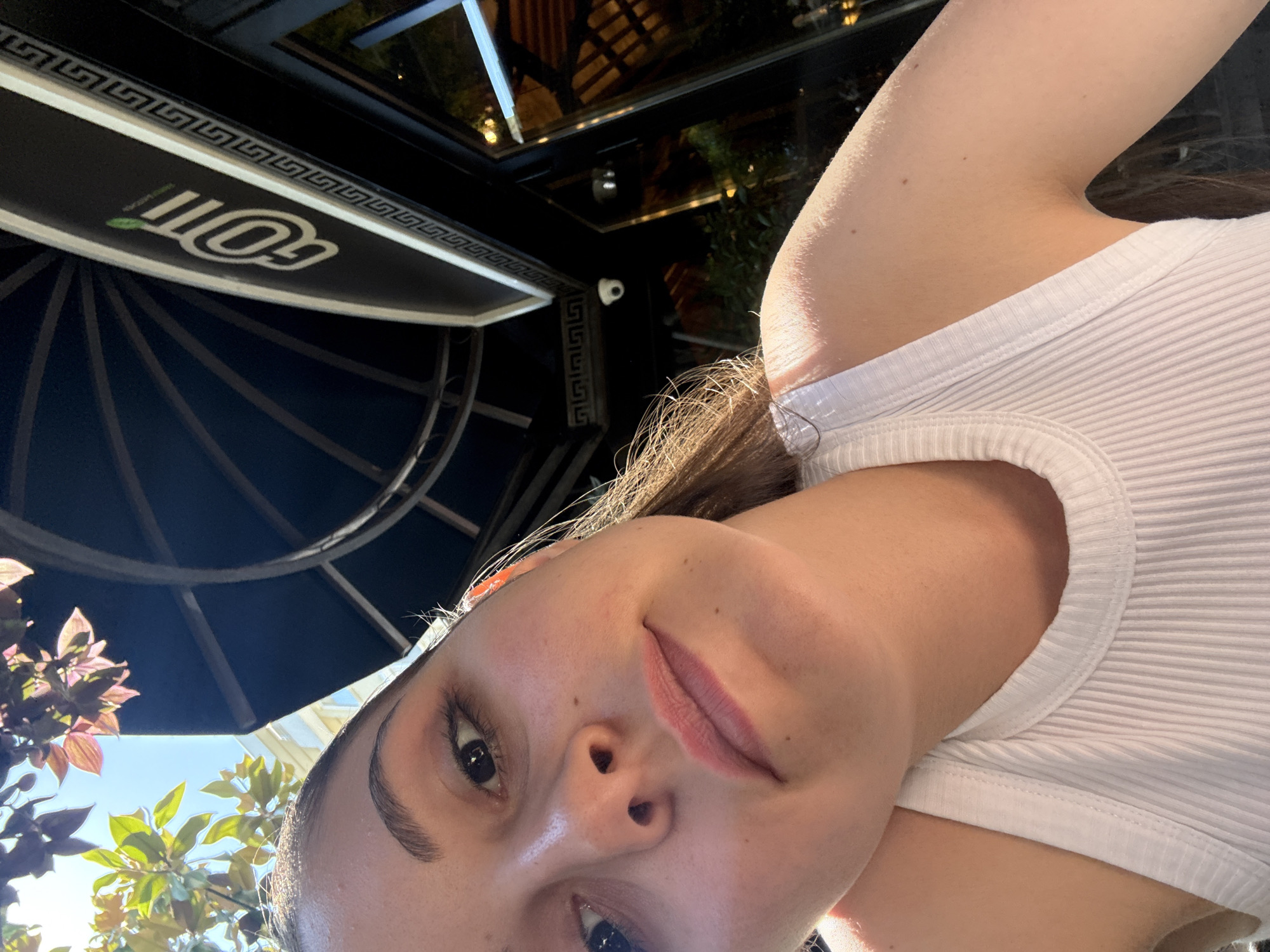 A close-up selfie-style shot captures a young woman looking directly at the camera, taken at what appears to be an outdoor cafe or restaurant in Burgas, Bulgaria. The image is rotated counter-clockwise. The woman's face dominates the foreground, featuring clear skin, dark eyes, and well-defined eyebrows. Her lips are slightly parted in a neutral to soft expression. She wears a simple white ribbed tank top, and her light brown hair frames her face.
In the background, a dark sign with a white border featuring a Greek key pattern is visible. Prominently displayed on the sign is the white text "GOTT," accompanied by a small green leaf-like logo and some smaller, indistinct text above it. This likely indicates "Gott Club & Grill," a known establishment in Burgas. Below the sign, a dark structure with radial spokes, possibly part of a large umbrella or awning, is seen. A small, white security camera is mounted on a column or beam in the mid-ground.
Above and behind the structures, a glass ceiling or canopy reflects glimpses of outdoor lights and trees. In the bottom left, against a bright blue sky, vibrant green foliage and some reddish-purple leaves add natural elements to the scene. The bright natural light suggests it is daytime, possibly late afternoon. The setting combines urban dining elements with natural light and plants.