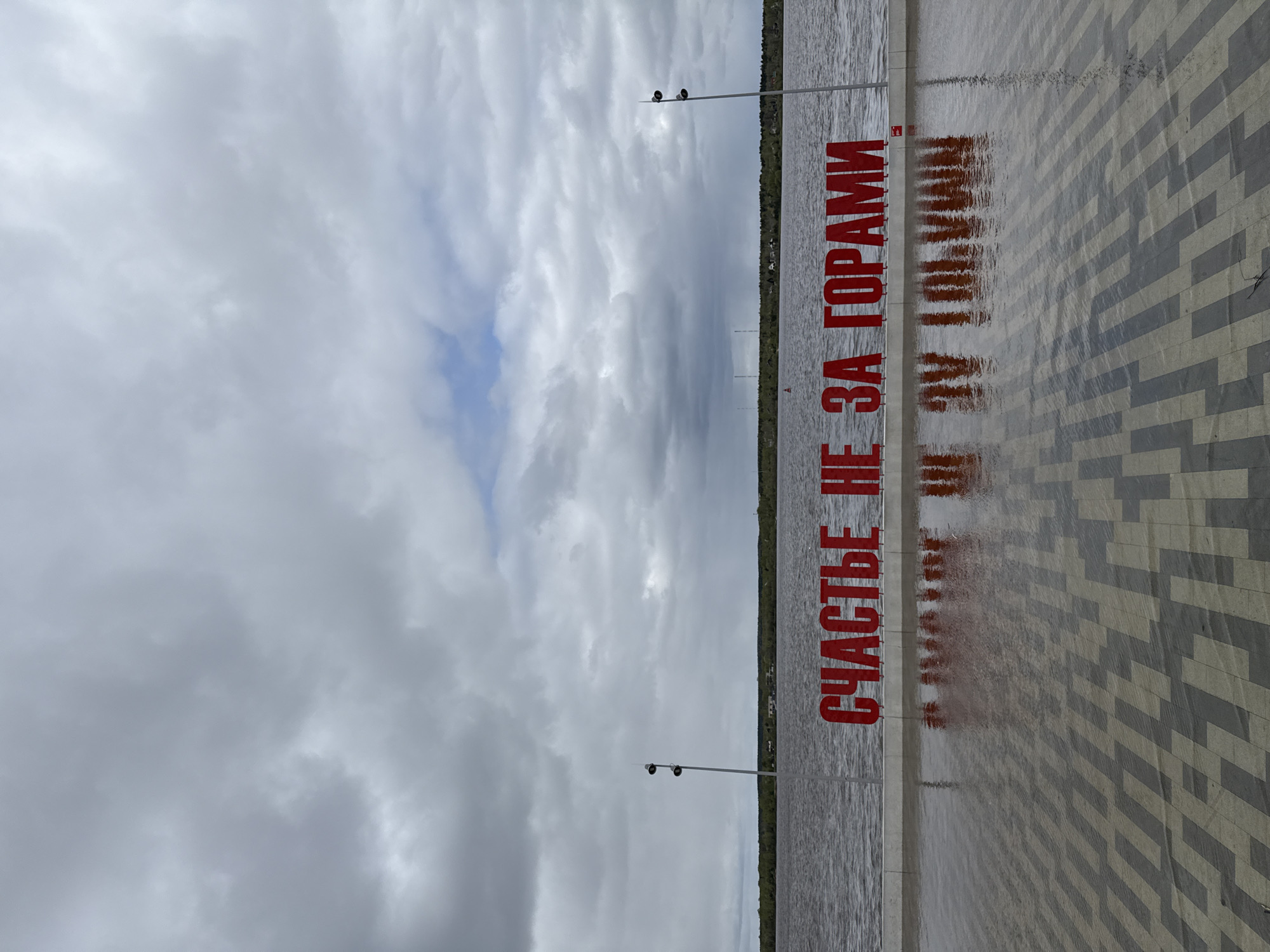 The image shows a view of the Kama River in Perm, Russia, with a large red sign that reads "Счастье не за горами" (Happiness is not beyond the mountains). The sign's reflection is visible on the wet pavement. The sky is overcast with clouds. A row of trees and poles with lamps or cameras are along the riverbank. The pavement is patterned with grey and white rectangular paving stones.