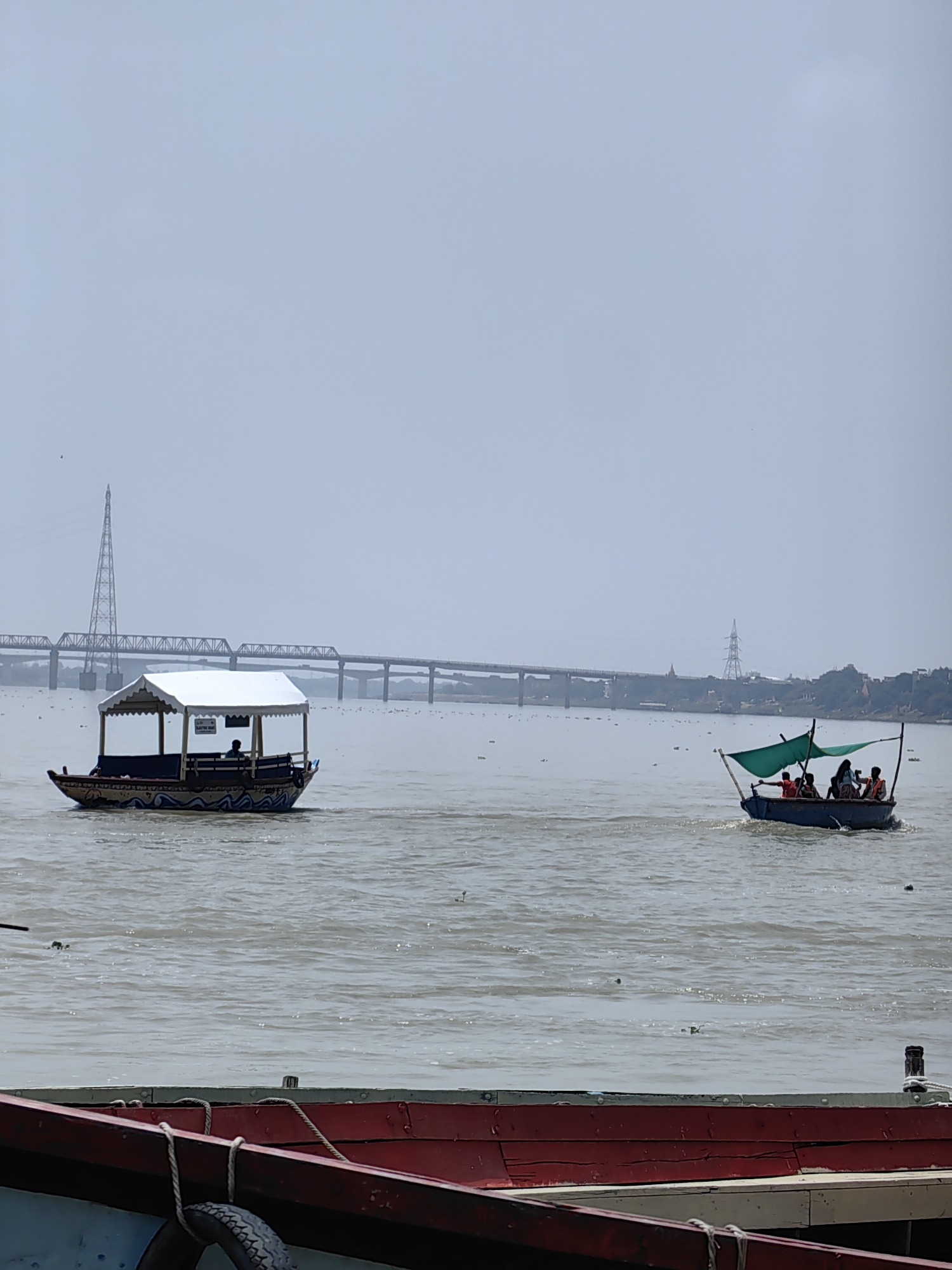 The image captures a wide shot of the Ganga river in Varanasi, India, on an overcast day. In the foreground, the red-painted edge of a docked boat is visible, with a spare tire hanging from its side. Further out on the river, two boats are in motion. The boat on the left is larger and covered with a white canopy, with a single person visible at the helm and what appears to be passengers seated. The boat on the right is smaller, with a green awning, carrying several passengers who are wearing orange life jackets. In the background, a long, multi-span bridge stretches across the river, with a prominent lattice tower visible to its left, likely a power transmission pylon. The water is a murky brown, with some debris scattered on its surface. The distant shoreline is lined with trees and structures, suggesting the presence of the city. The overall atmosphere is hazy, typical of a monsoon or polluted environment.