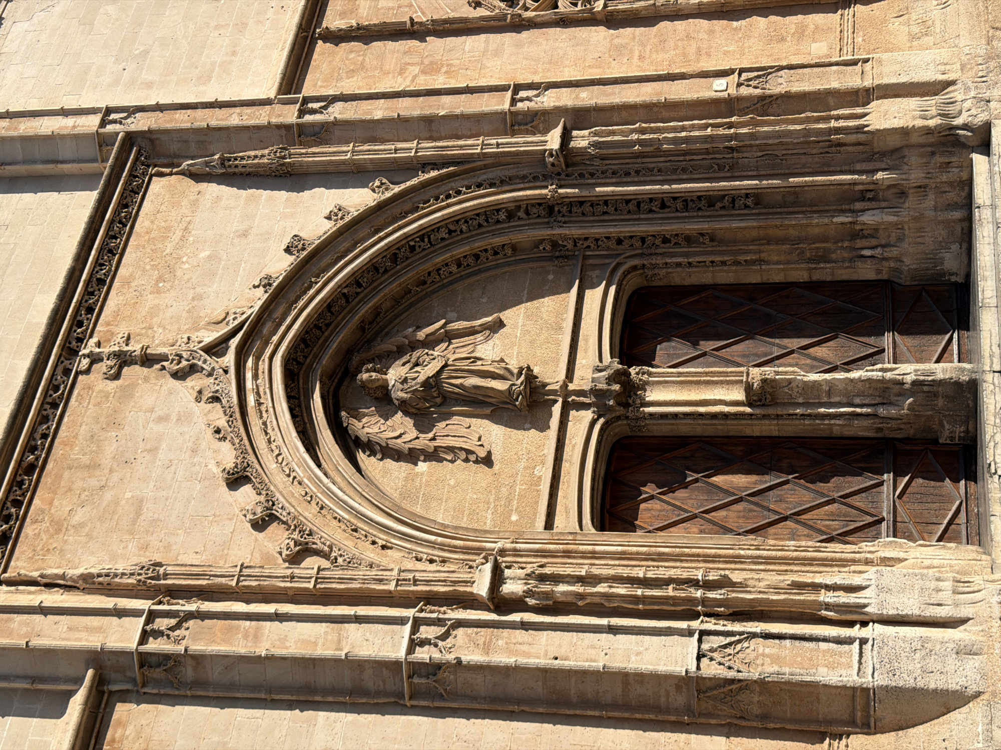 The image shows a close-up of an ornate stone doorway of a building in Palma de Mallorca, Spain. The doorway is arched and features intricate carvings, including a sculpture of an angel above the double wooden doors. The doors are made of dark wood with a diamond pattern. The stonework is weathered and appears to be old. The overall impression is one of historical and architectural significance.