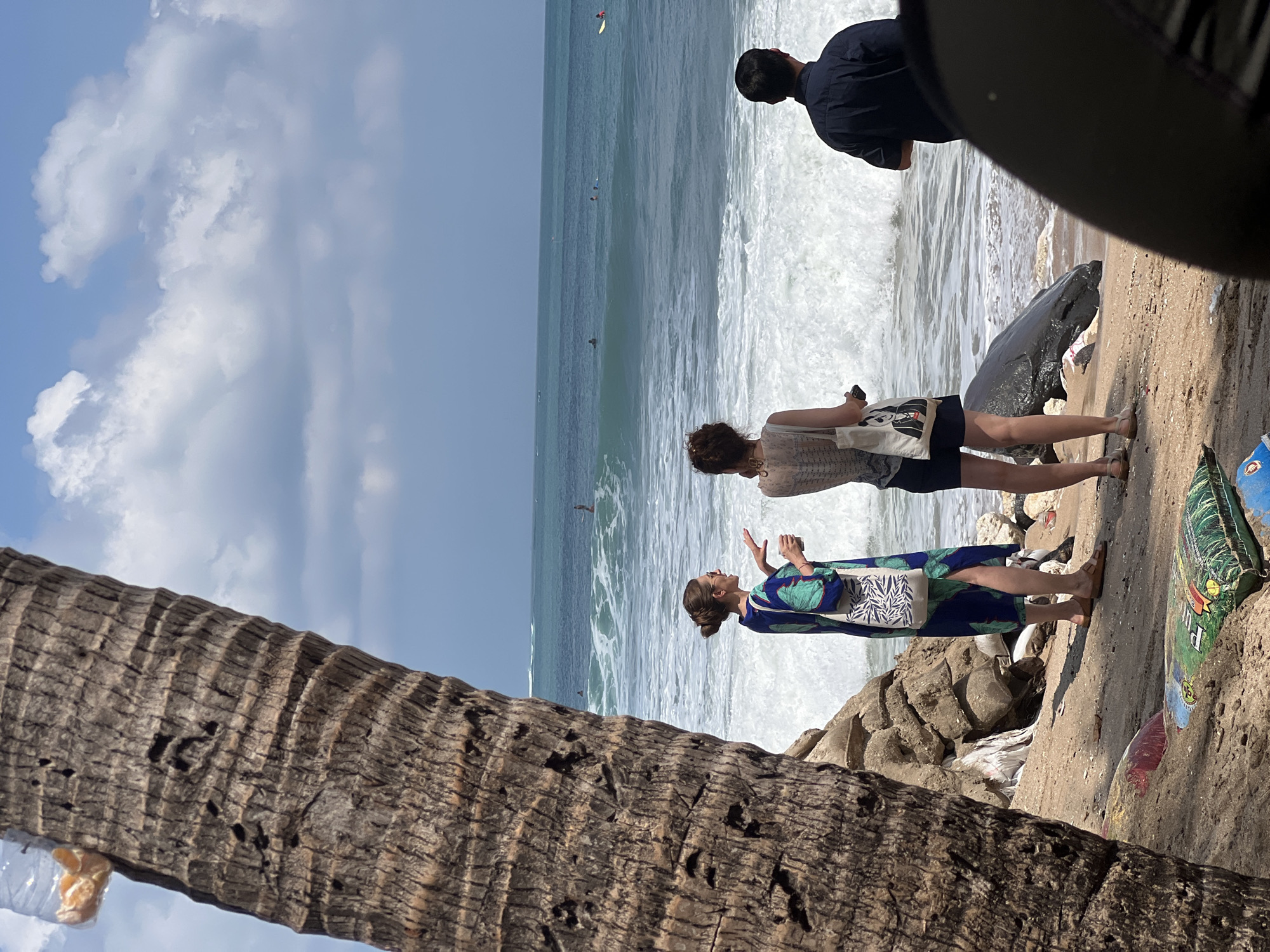 In Kuta, Indonesia, on a sun-drenched beach, a group of people stand near the shoreline. A large palm tree trunk, weathered and textured, dominates the left foreground, partially obscuring the view. Behind it, two women are engaged in conversation. One woman, wearing a blue and green patterned dress and sandals, holds a cup and gestures with her hand. She carries a tote bag with a black and white leaf pattern. The other woman, standing to her right, wears a light-colored top with a patterned strap, dark shorts, and sandals, and holds a phone or small device. To their right, a man in a dark blue shirt stands facing the ocean.
The sea displays a turquoise hue under a sky dotted with fluffy white clouds. Waves crash onto the sandy shore, creating a line of white foam. Several surfers are visible in the distance on the water. A large dark, possibly wet, object, resembling a fallen log or debris, lies on the sand near the water's edge. The beach itself shows scattered debris, including colorful plastic items and what appear to be sandbags. The overall scene captures a moment of coastal life, with a mix of leisure and natural elements.