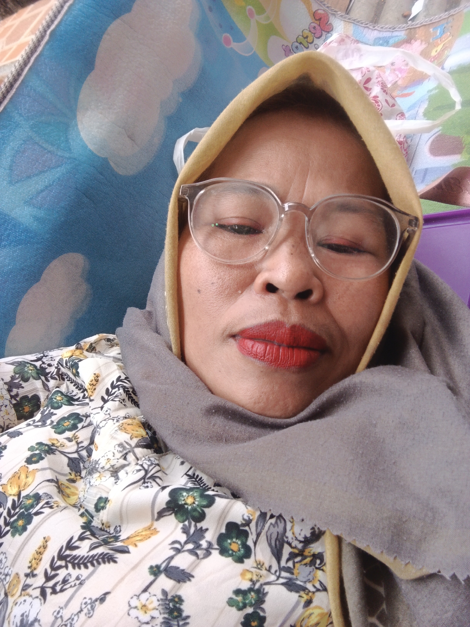 A close-up, selfie-style portrait of an adult Indonesian woman, likely in Warungkiara, Indonesia, shows her head and shoulders. She is wearing a yellow headscarf, a grey scarf draped around her neck, and a floral print blouse. She has on clear-rimmed glasses and bright red lipstick. Her eyes are partially closed. The background is out of focus, but appears to be a colorful, possibly fabric-covered surface with a blue patterned section. The lighting suggests an indoor setting, possibly a home.