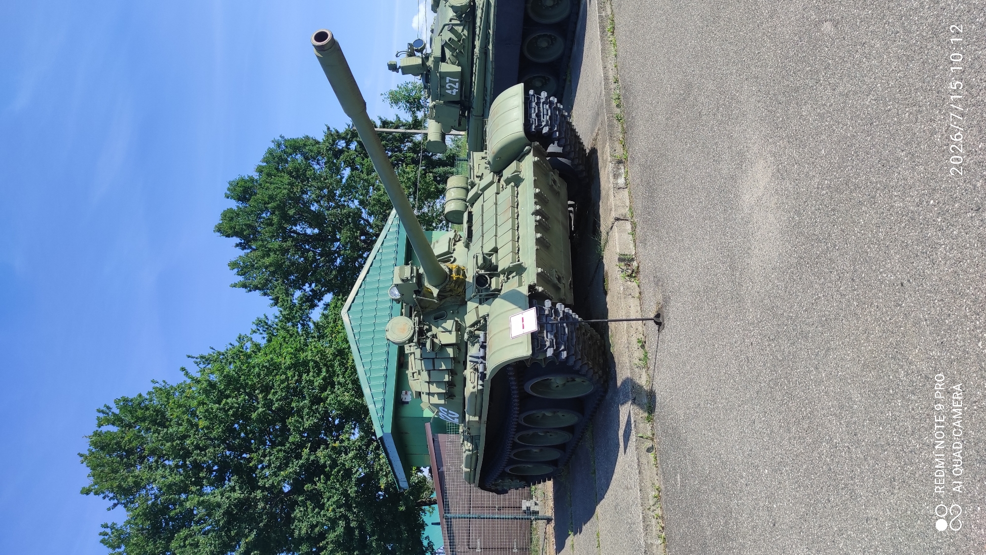 A matte olive-green military tank, identified as a T-54/55 variant, is parked on a concrete pad in front of a low, green building with a pitched roof. The tank is facing towards the right of the frame and features a long main gun barrel extending forward and slightly upwards. It has a multi-wheeled tread system and various components typical of a tank, including a turret with what appears to be a machine gun mount. A small white sign with a red stripe is positioned in front of the tank. In the background, lush green trees with dense foliage are visible, and the sky is a clear, bright blue with a few wispy clouds. To the right of the primary tank, another, partially visible tank is present, displaying the number "427" on its turret. The scene is well-lit by sunlight, casting shadows on the ground. The overall setting appears to be an outdoor exhibit or museum display. The image also contains overlaid text in the bottom left and right corners, indicating "REDMI NOTE 9 PRO AI QUAD CAMERA" and "2026/7/15 10:12" respectively, suggesting it is a photograph taken with a mobile device. The presence of a fence to the left of the tank implies a perimeter or enclosure. The location is identified as Лашанскі сельскі Савет, Belarus, implying an outdoor setting within that administrative region.
