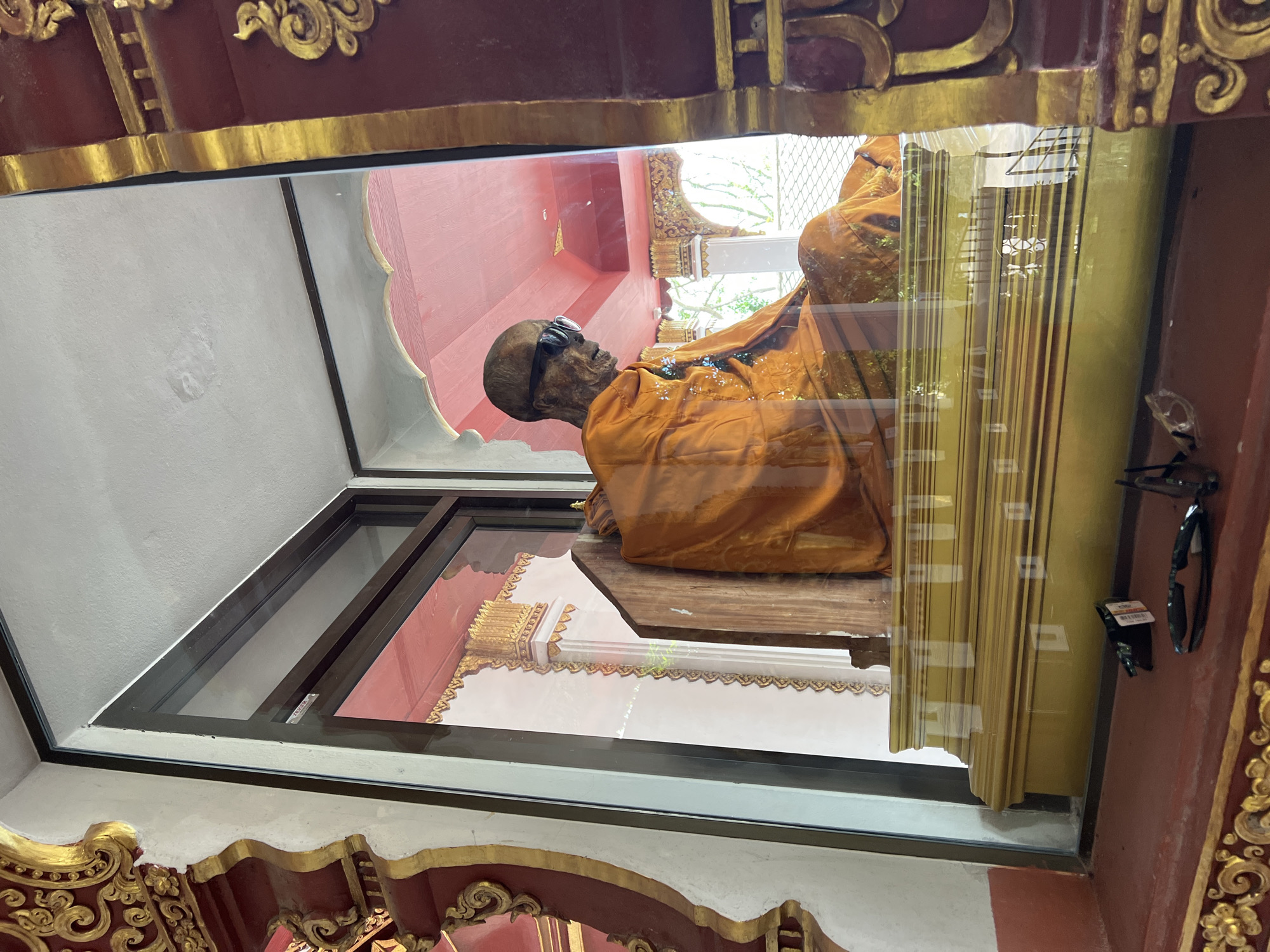 A mummified monk wearing sunglasses and an orange robe sits cross-legged on a golden platform inside a glass display case in Baan Hua Thanon, Thailand. The monk's body appears withered and preserved. The display is situated within a richly decorated building with red and gold accents. In front of the glass case, on a red ledge, are a pair of sunglasses and some other small objects. The background visible through the display case shows architectural details of a temple or shrine, including ornate pillars and a red ceiling. Sunlight reflects off the glass, obscuring some details of the scene.
