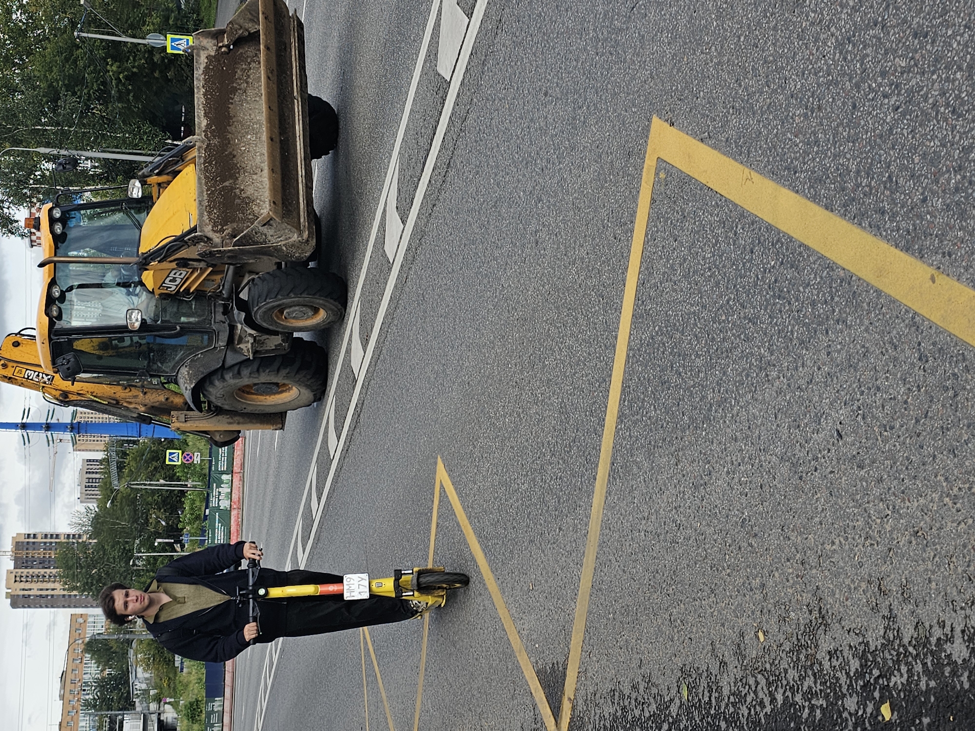 A young man rides a yellow electric scooter down a city street in Moscow, Russia. He is wearing a dark hooded jacket over a khaki polo shirt and dark pants. He has earbuds in and is looking towards the camera. The scooter has a license plate that reads "HM9 17Y". Behind him, a yellow JCB backhoe loader drives down the road, its bucket filled with dirt. The road has yellow and white markings. In the background, tall apartment buildings and trees are visible. The sky is overcast.