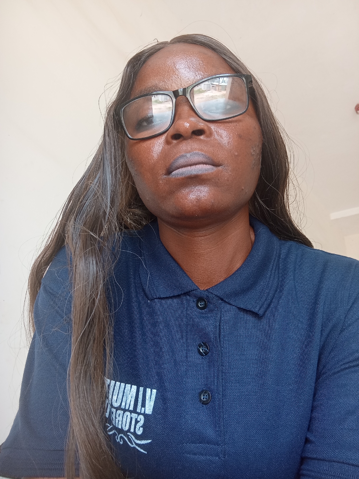 A close-up, low-angle, medium shot captures a dark-skinned woman wearing glasses and a dark blue collared shirt. Her long, dark hair is parted on the left and falls over her right shoulder. She is wearing dark lipstick. The background is a plain, light-colored wall. A white logo with stylized text and wavy lines is visible on the front of her shirt. The lighting is bright and appears to be from an overhead source.