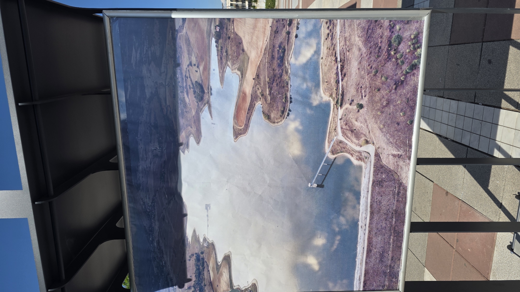 The image depicts a framed aerial photograph of a landscape featuring a lake and surrounding terrain. The photo, mounted in a public display structure, shows the body of water with an irregular shoreline bordered by fields and vegetation. A causeway or pier extends into the lake. The photograph's surface exhibits some glare and possible reflections. The display structure is positioned on a paved area constructed from various colored bricks. The image was taken in Blagoevgrad, Bulgaria.