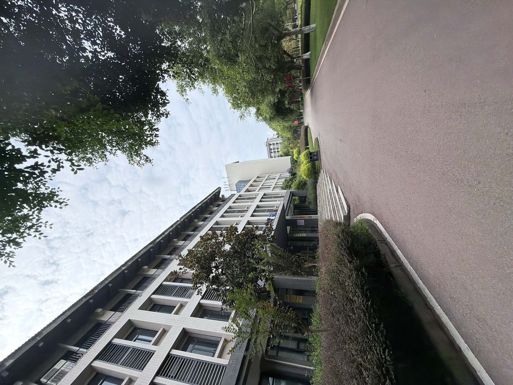 In the city of Jiangjin, China, a wide, paved pathway curves through a lush green environment. The pathway is bordered on the left by a dense, dark green hedge and a light-colored building with multiple floors. The building features a rhythmic pattern of windows with dark louvers, and balconies with black railings. Several trees with dark green foliage are planted in front of the building. On the right side of the pathway, a manicured lawn is dotted with mature trees, some of which have their trunks painted white. In the distance, additional buildings are visible, and the sky is partly cloudy. The scene captures a tranquil, campus-like setting.
