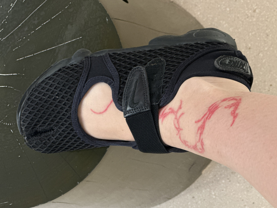 A high-angle, close-up photograph depicts a light-skinned human left foot. The foot is clad in a black Nike Air Rift shoe, characterized by its split-toe design, mesh upper, and two black hook-and-loop straps; the upper strap features a white Nike swoosh logo. Red, temporary markings are drawn on the skin of the ankle and upper foot. A crescent shape is visible on the upper ankle, while a larger, more abstract, stylized design, resembling a branching or avian form, covers the lower ankle and dorsum of the foot. The foot is positioned above a dark, cylindrical object, possibly a bench or planter, which exhibits several light-colored linear scuff marks on its surface. A light-colored, flat surface, consistent with pavement, is partially visible to the left of the foot. No specific architectural features or landmarks are present in the image to identify a precise location within the city of Paris, France.