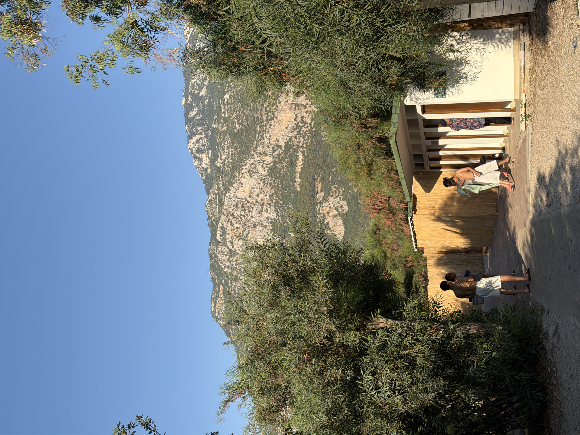 This image captures a scene at a beachside facility with changing rooms in Fethiye, Turkey. In the foreground, three young men are visible. One man, wearing white shorts, stands facing away from the camera, with his back to a light-colored building with multiple doorways. To his left, another man, partially obscured, appears to be holding a towel. Further to the right, a third man is exiting one of the changing rooms, holding a towel. The changing room structure has a light-colored facade with wooden doors and a green roof. Behind the building, a tall fence constructed of woven reeds runs horizontally across the frame, casting long shadows from the sunlight. The ground is covered in a mixture of gravel and sand. Lush green trees with thin leaves are prominent on both the left and right sides of the image, framing the scene. In the background, a large, rugged mountain dominates the skyline, with its slopes covered in sparse vegetation and rocky outcrops. The sky is clear and a vibrant blue. A paraglider is visible as a small dot in the upper portion of the mountain.