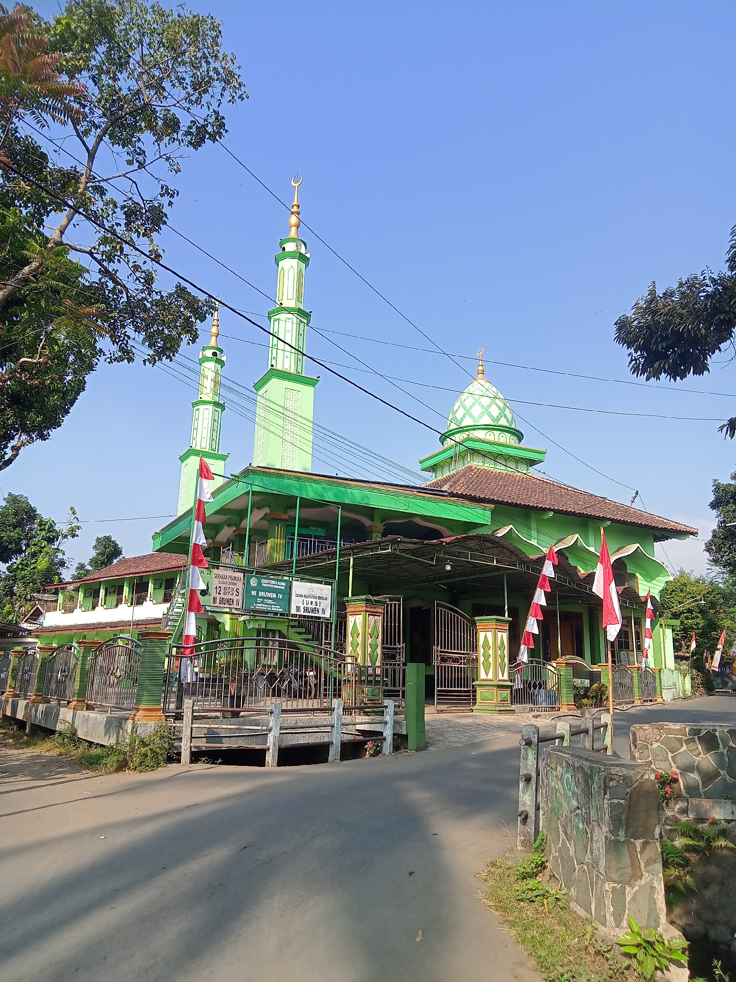 The image depicts the exterior of a bright green mosque in Sruwen, Indonesia, bathed in clear blue sky and sunlight. Two tall, slender minarets with golden crescents stand prominently, flanking the main building. The mosque features a traditional tiled roof and a green, patterned dome. Red and white Indonesian flags are displayed on poles along the front of the building, suggesting a national celebration or observance. The mosque appears to be situated near a road, with a small concrete bridge crossing a channel or ditch in the foreground. Trees with green foliage surround the structure, and power lines are visible in the sky. Signs in Indonesian are attached to the mosque, one indicating it is part of the "KEMENTERIAN AGAMA KABUPATEN SEMARANG" (Ministry of Religious Affairs, Semarang Regency) and another that appears to be related to a school, "MI SRUWEN IV". There are no people clearly visible in the image.