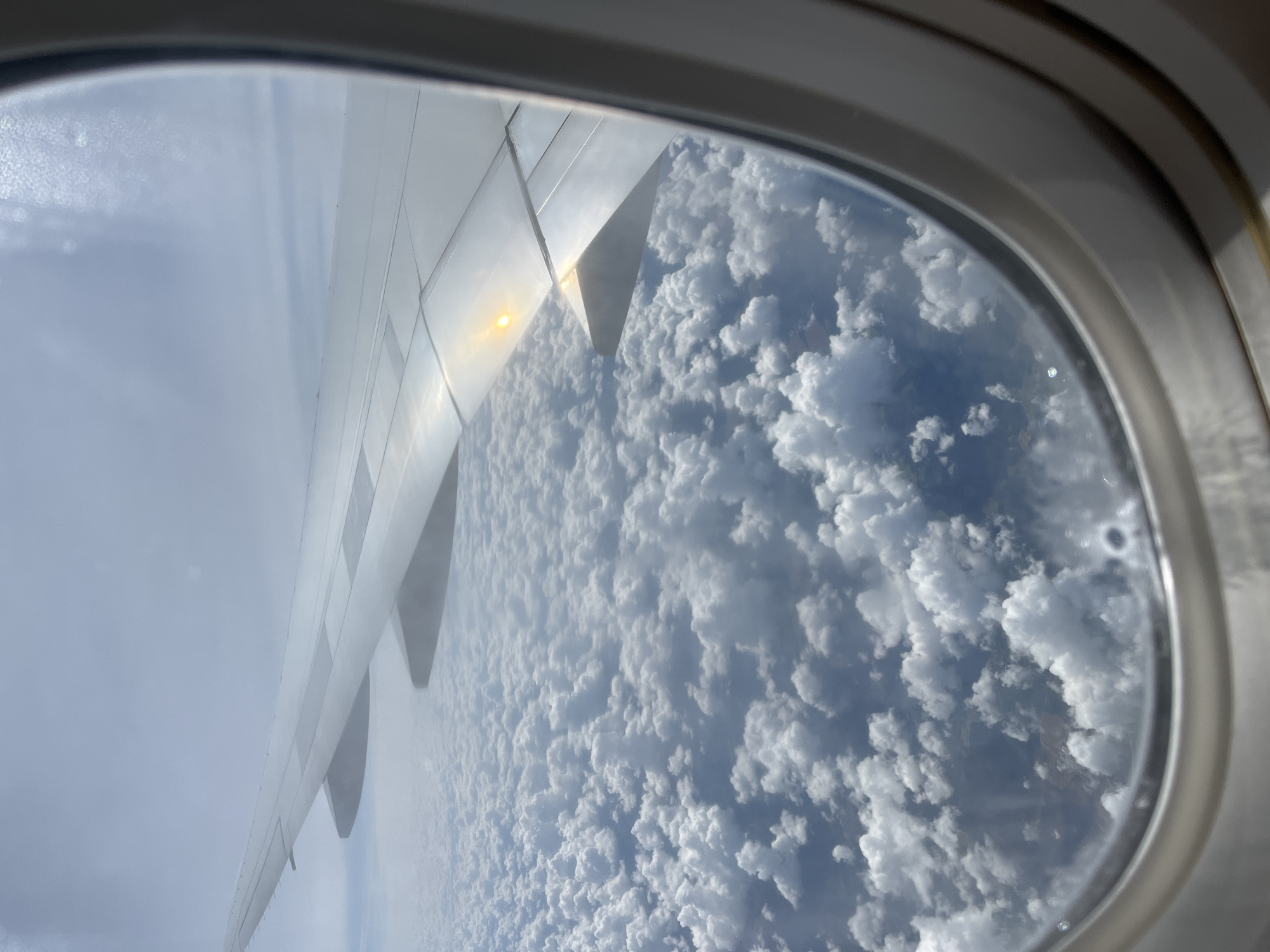 An aerial view from an airplane window shows a vast expanse of fluffy white clouds below. The wing of the airplane is visible in the foreground, extending across the frame from left to right. Sunlight glints off the surface of the wing, creating a bright yellow reflection. The clouds appear dense and cover the entire landscape beneath. The image is taken from a high altitude, suggesting the airplane is in flight. There are no visible signs of the city of Sukhanikha, Russia, or any urban features in this image.
