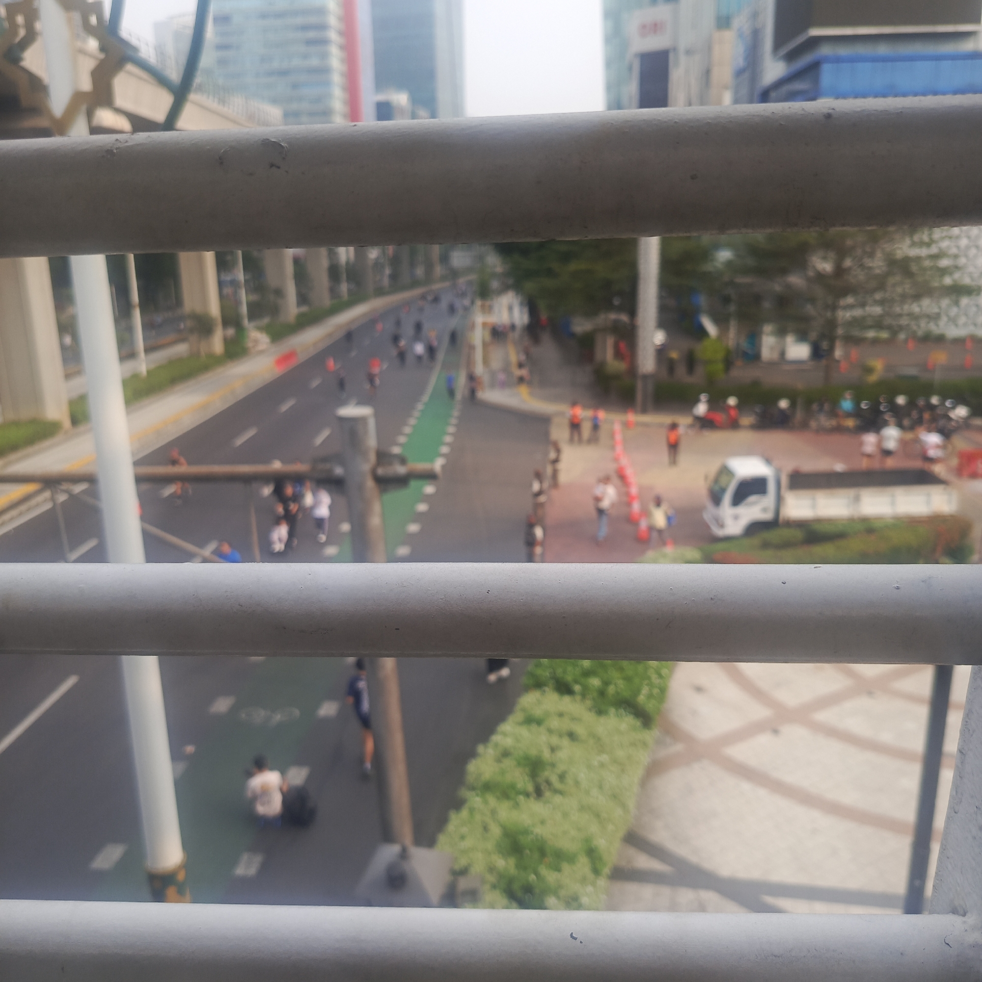 The image provides an elevated, blurred view of a street scene in South Jakarta, Indonesia, likely during an event. In the foreground, two horizontal grey railings partially obscure the view. Below the railings, a multi-lane road is visible, with traffic cones and a green lane marking indicating a closed or restricted area. Several individuals are present on the road and sidewalk, some appearing to be walking or running. On the road, there are a few individuals scattered, and what looks like a white truck with a black bed is parked on the right side of the road. The background features tall modern buildings, characteristic of a city environment. The overall impression is one of an urban event or activity taking place on a street that has been modified for the occasion, with people participating and city infrastructure in the backdrop.